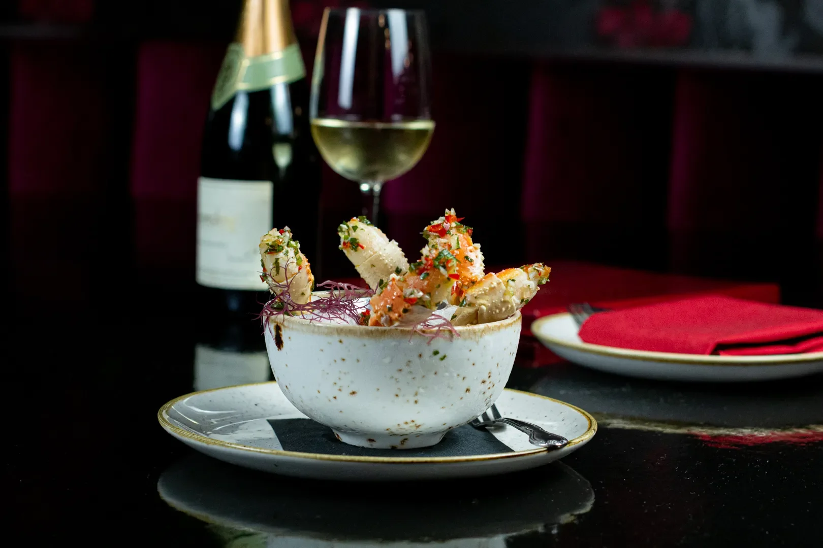 A bowl of stuffed artichoke hearts garnished with chopped herbs, placed on a plate with a fork, alongside a bottle of champagne and a glass of white wine in the background, on a dark table with red napkins.
