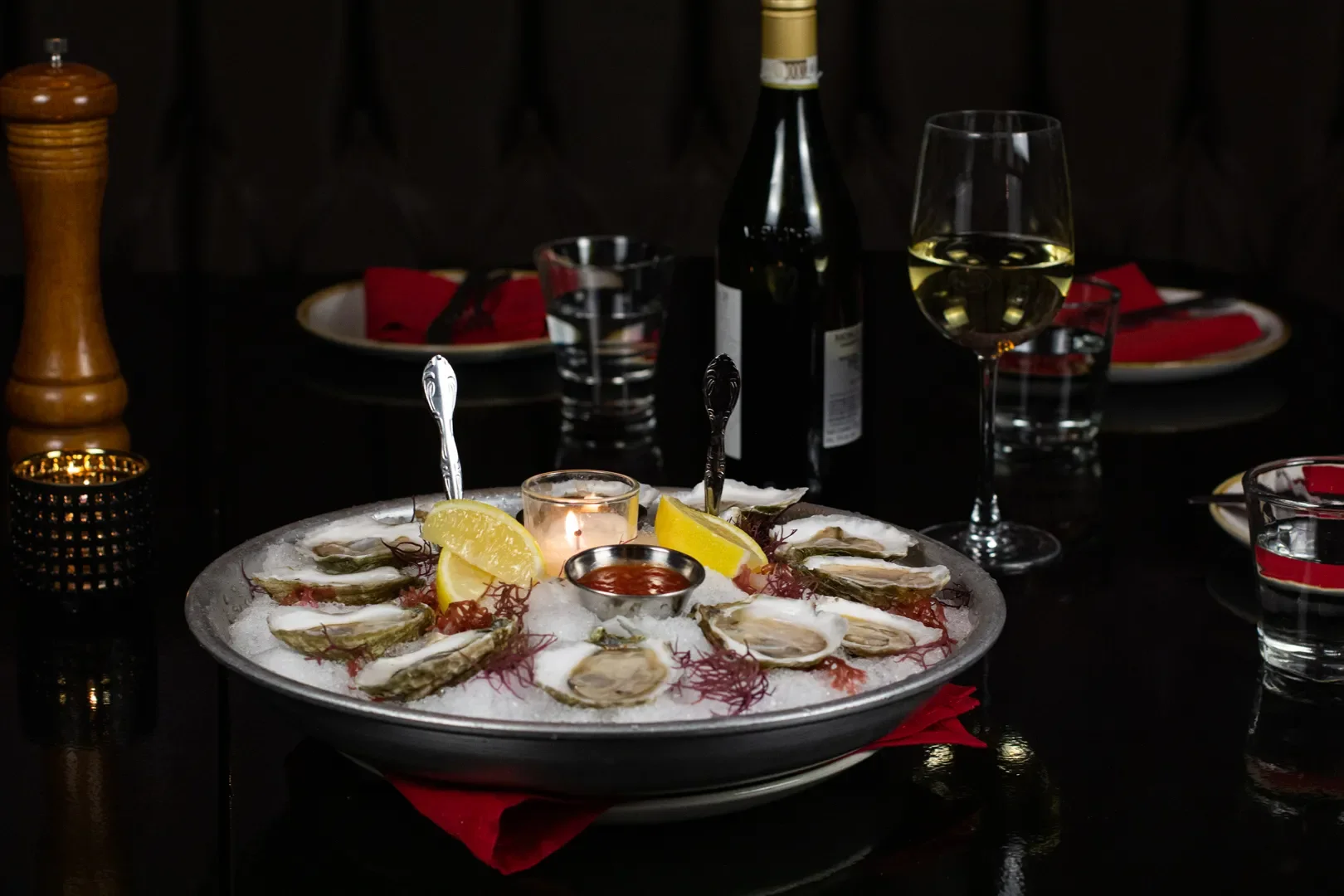 A seafood platter with oysters on the half shell, lemon wedges, and a small cup of cocktail sauce on a bed of crushed ice, accompanied by a lit candle in the center, on a dark table set for dinner.