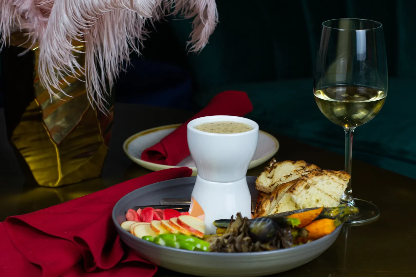 A dinner table setting with a plate of assorted vegetables, bread, and a white sauce, a glass of white wine, and a cup of soup, on a dark surface with a red napkin and a gold vase in the background.