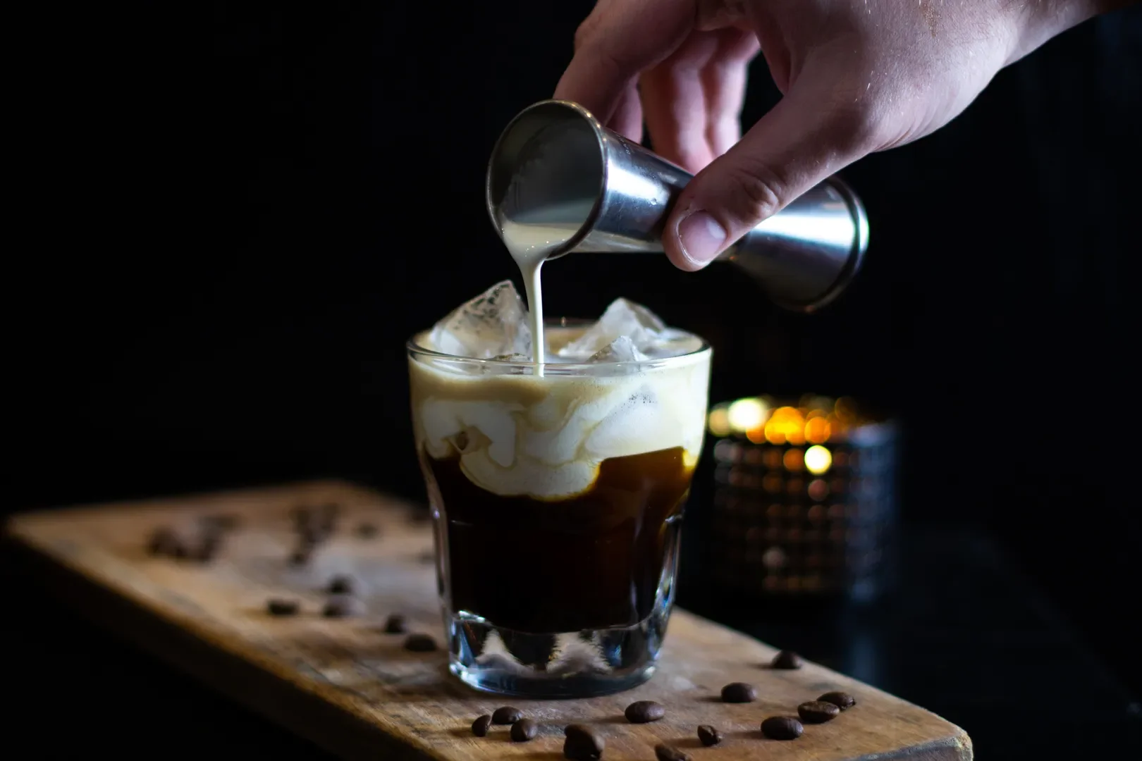 A person pouring creamer into a glass of iced coffee with whipped cream, sitting on a wooden board with coffee beans around it.