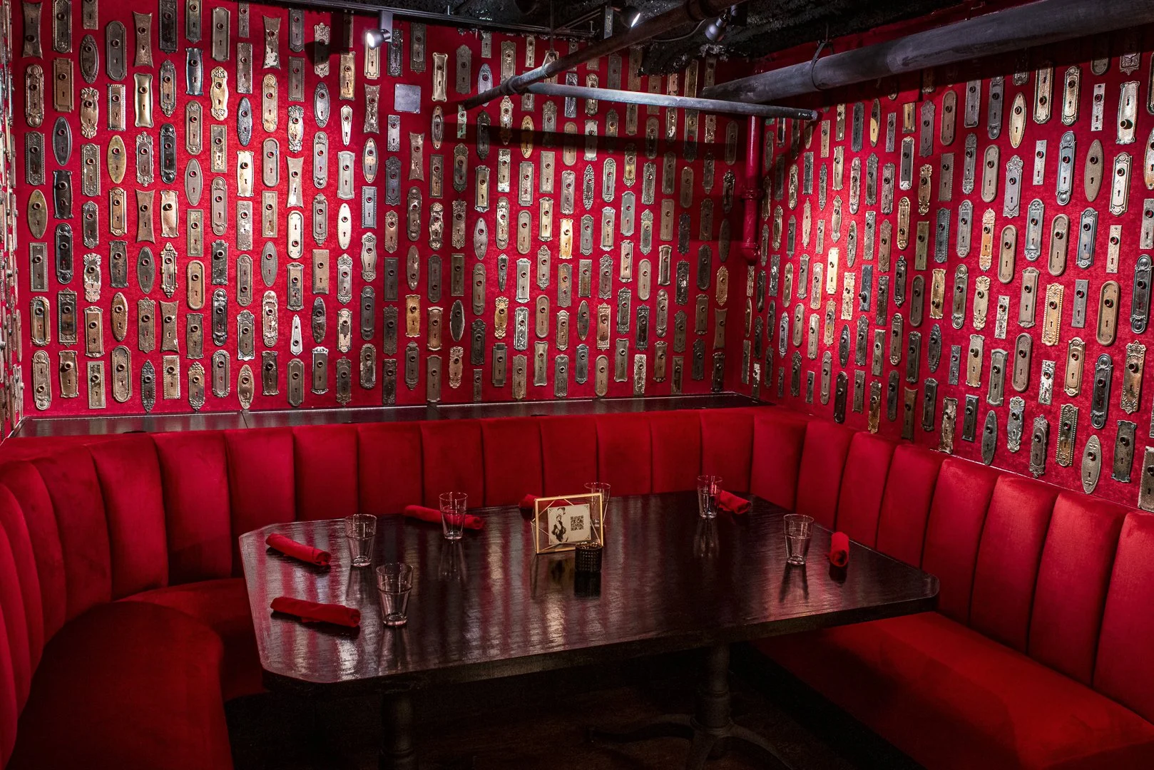 A cozy restaurant corner with a red upholstered banquette, dark wooden table, glasses, red napkins, and a small framed menu or sign. The background wall features a red patterned wallpaper with various vintage keys.