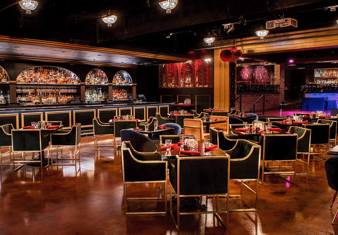 Interior view of an elegant bar and restaurant with black and gold furniture, red napkins on tables, a well-stocked bar with bottles, and warm ambient lighting.