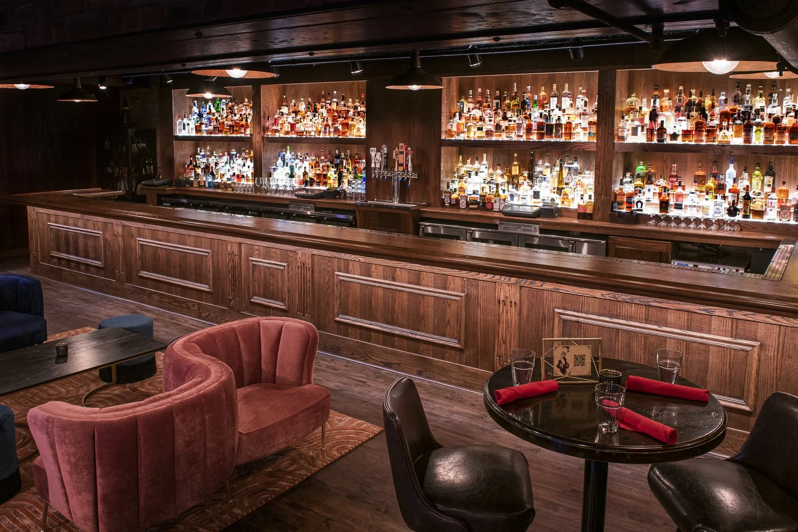 Interior of a bar with a long wooden counter, illuminated shelves filled with various bottles of alcohol, and a seating area with pink velvet chairs, black chairs, and a round table with red napkins and glasses.