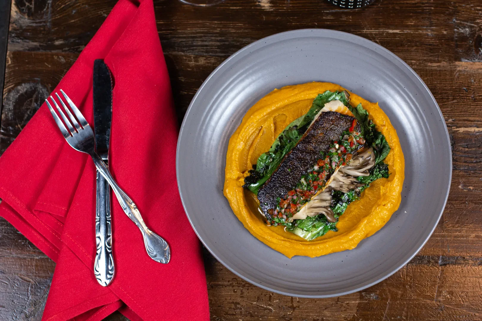 A plate of food with a piece of cooked fish topped with herbs and vegetables, served over a swirl of yellow puree, garnished with greens, on a gray ceramic plate. A red cloth napkin with a fork and knife arranged on it is placed next to the plate on 