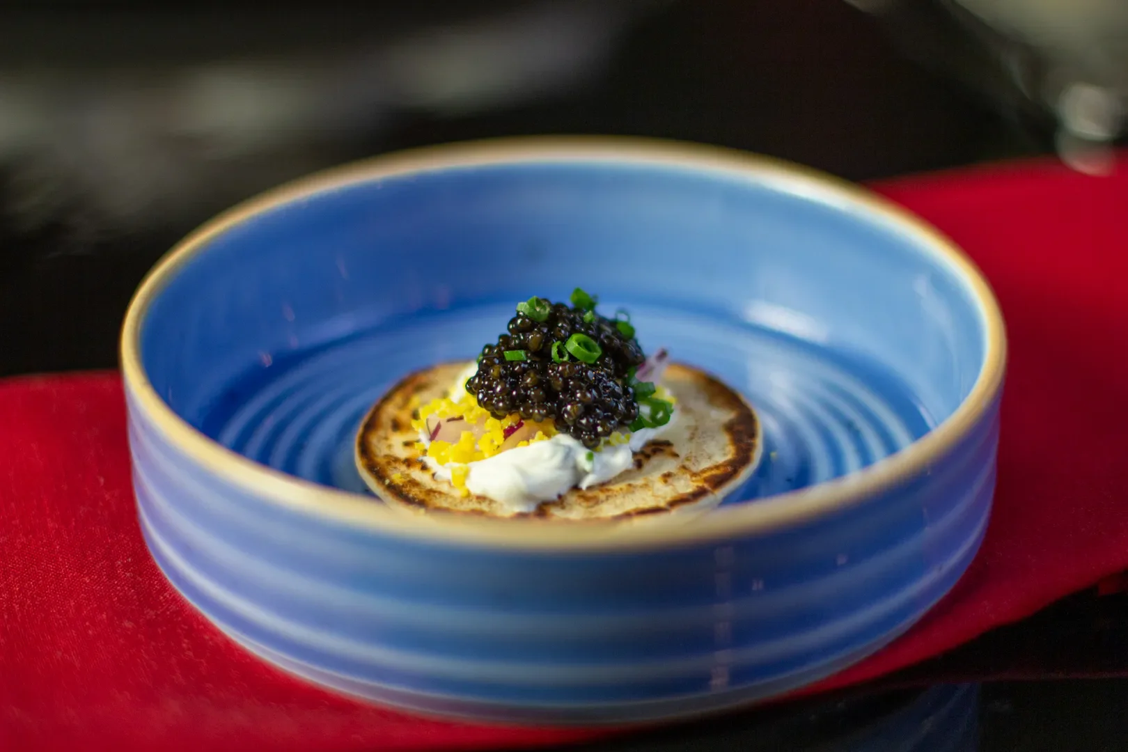 A blue ceramic bowl with a single mini pancake topped with black caviar, chopped green onions, and a dollop of sour cream, resting on a red cloth.