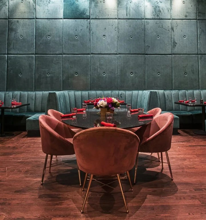 Elegant dining area with a round black table set with red napkins, glasses, and a floral centerpiece, surrounded by pink velvet chairs and a green cushioned banquette against a textured teal wall.
