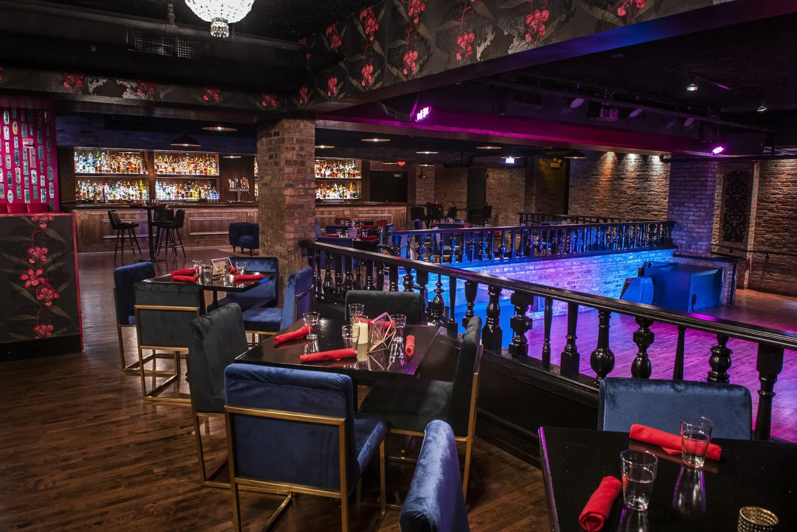 An empty bar and dining area with tables set with glasses, tissues, and red napkins, velvet chairs, exposed brick walls, a staircase with a black balustrade, and a colorful bar shelf illuminated with neon lighting.