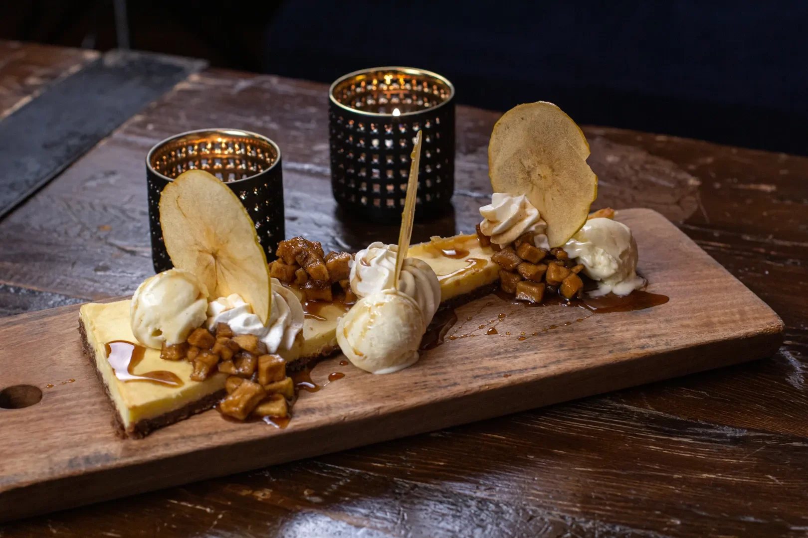 Banana split dessert on a wooden board featuring sliced bananas, vanilla ice cream scoops, whipped cream, caramel sauce, chopped nuts, and thin apple slices, with two black candle holders in the background.