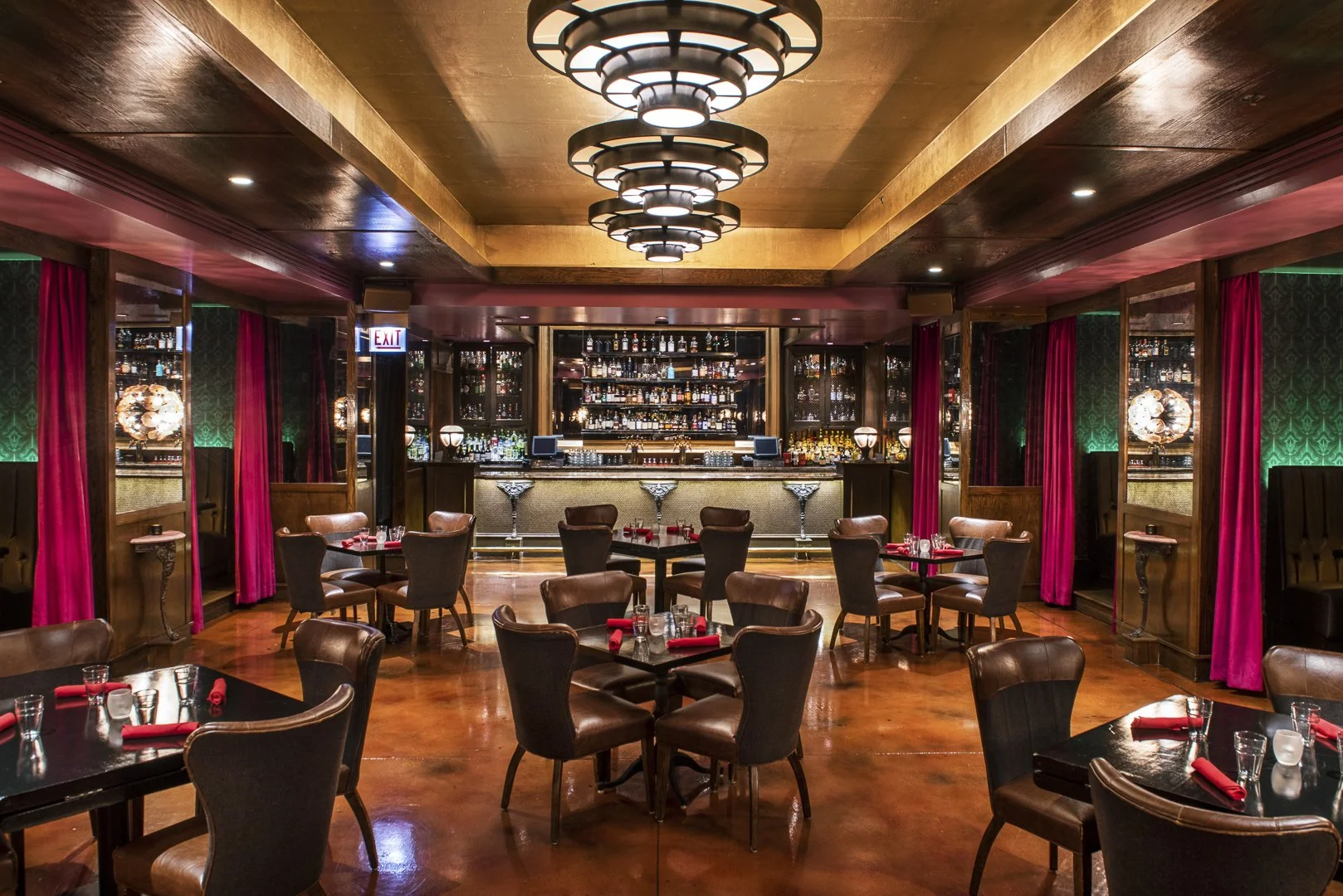 Empty upscale restaurant or bar with round tables set with glasses, napkins, and utensils, wooden floors, and a bar area with shelves of liquor bottles in the background, illuminated by ceiling and wall lighting, with red curtains and green wall panels.
