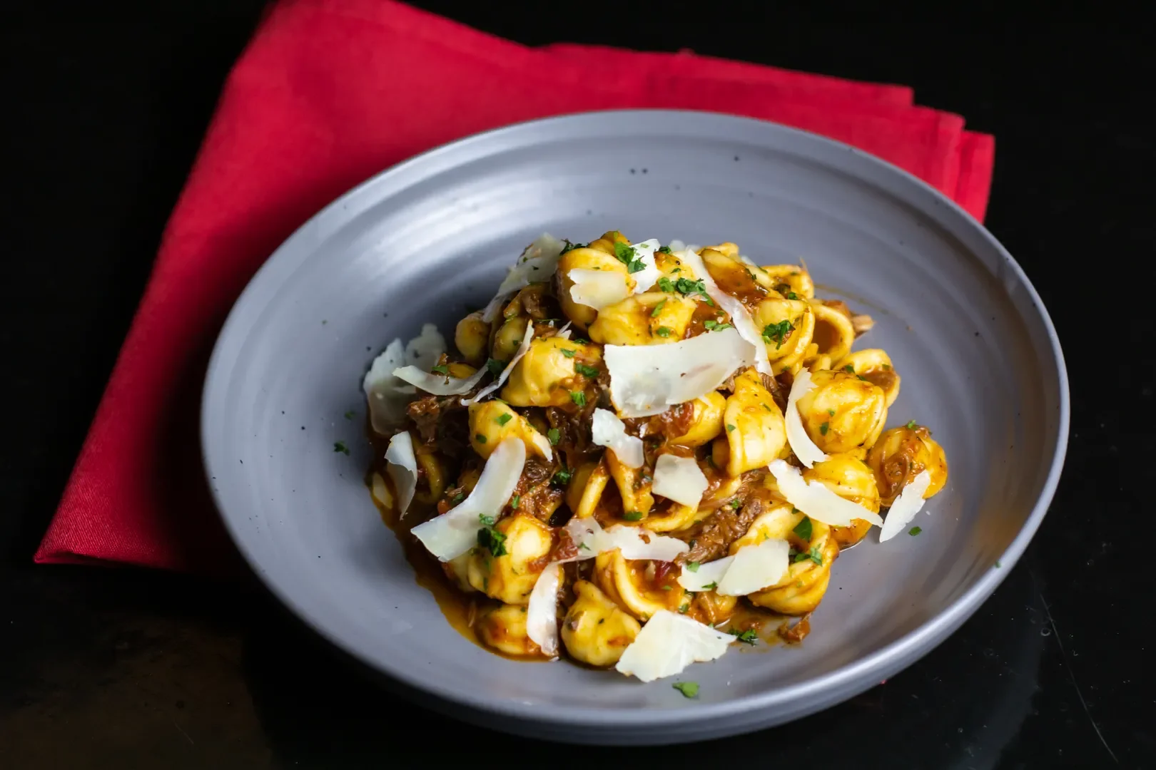 A plate of pasta with cheese shavings, garnished with chopped herbs, on a dark surface with a red napkin underneath.