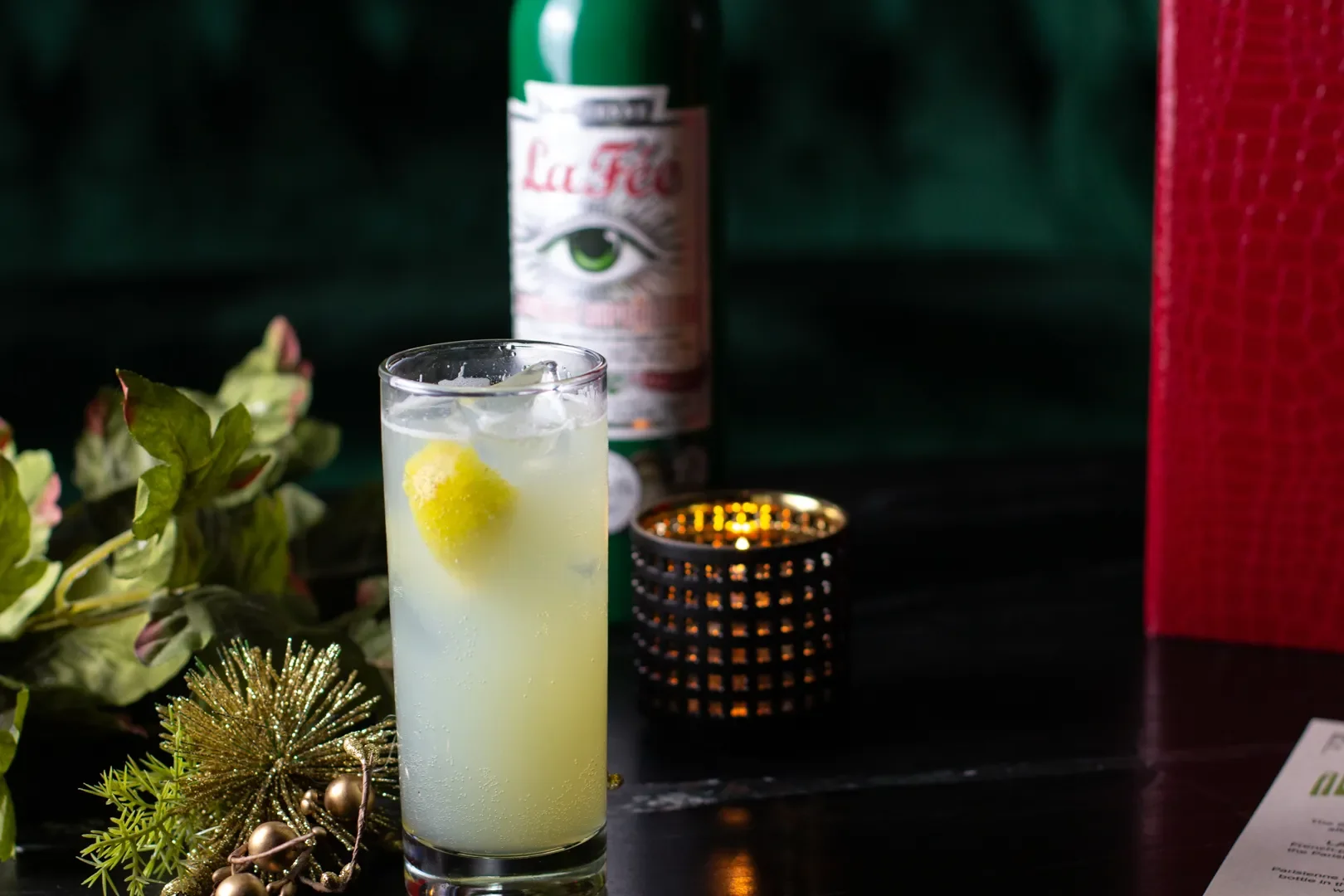 A tall glass filled with a yellow-green beverage, ice cubes, and a lemon slice, placed on a table with holiday decorations including a gold and green ornament, a green plant, and a red gift box. A green bottle with a label and eye illustration is in 