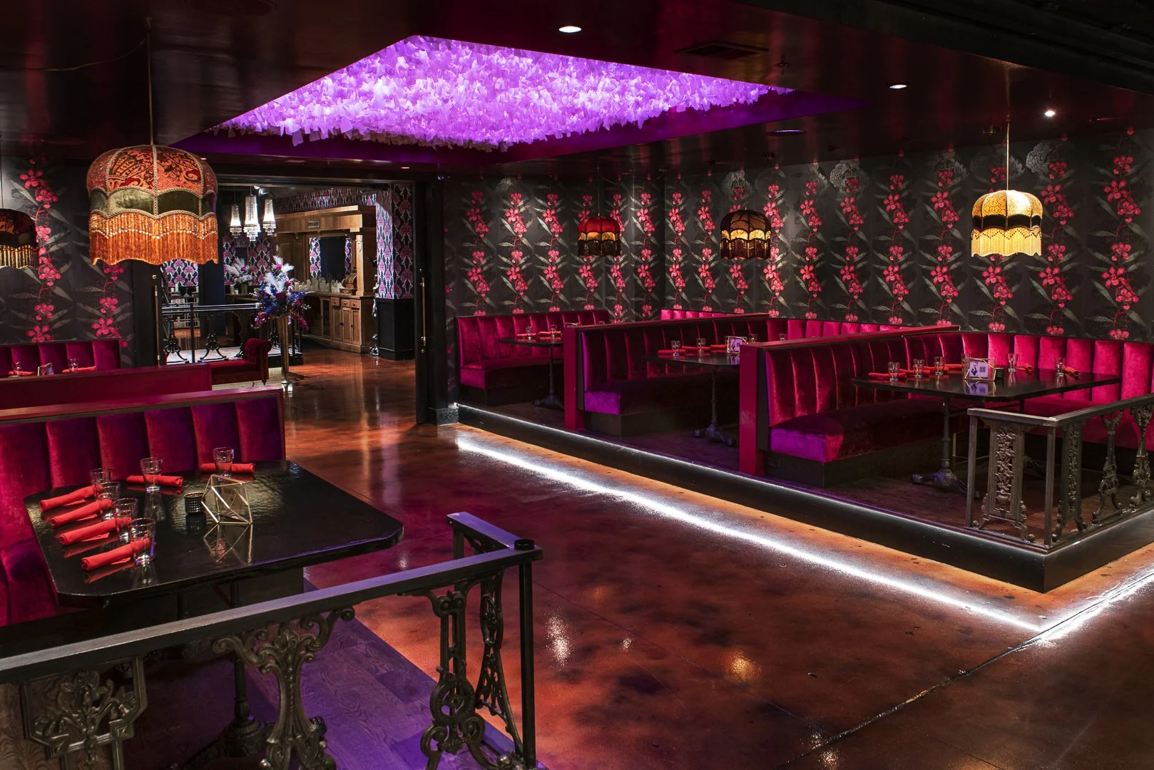 Interior of a restaurant with red velvet booths, dark floral wallpaper, and vintage hanging lamps with fringes, illuminated by pink and warm lighting.