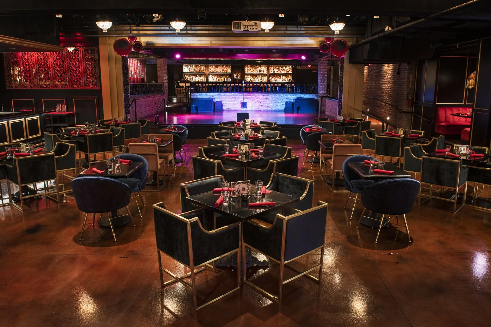 Empty stage with microphone at center, surrounded by seating area with tables and chairs set for dining, in a nightclub or entertainment venue with dim lighting, red and purple ambiance, and a bar at the back.