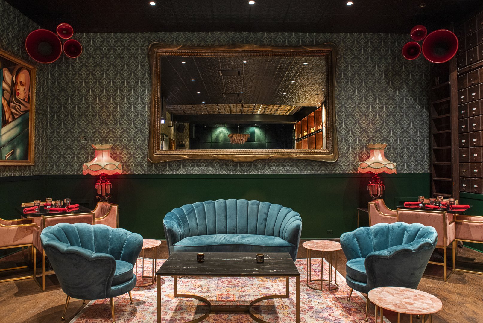 A stylish lounge with teal velvet armchairs and a matching sofa centered around a black coffee table. Behind the seating, there are pink velvet side chairs at a table with red napkins and glasses, illuminated by two ornate table lamps with pink fring