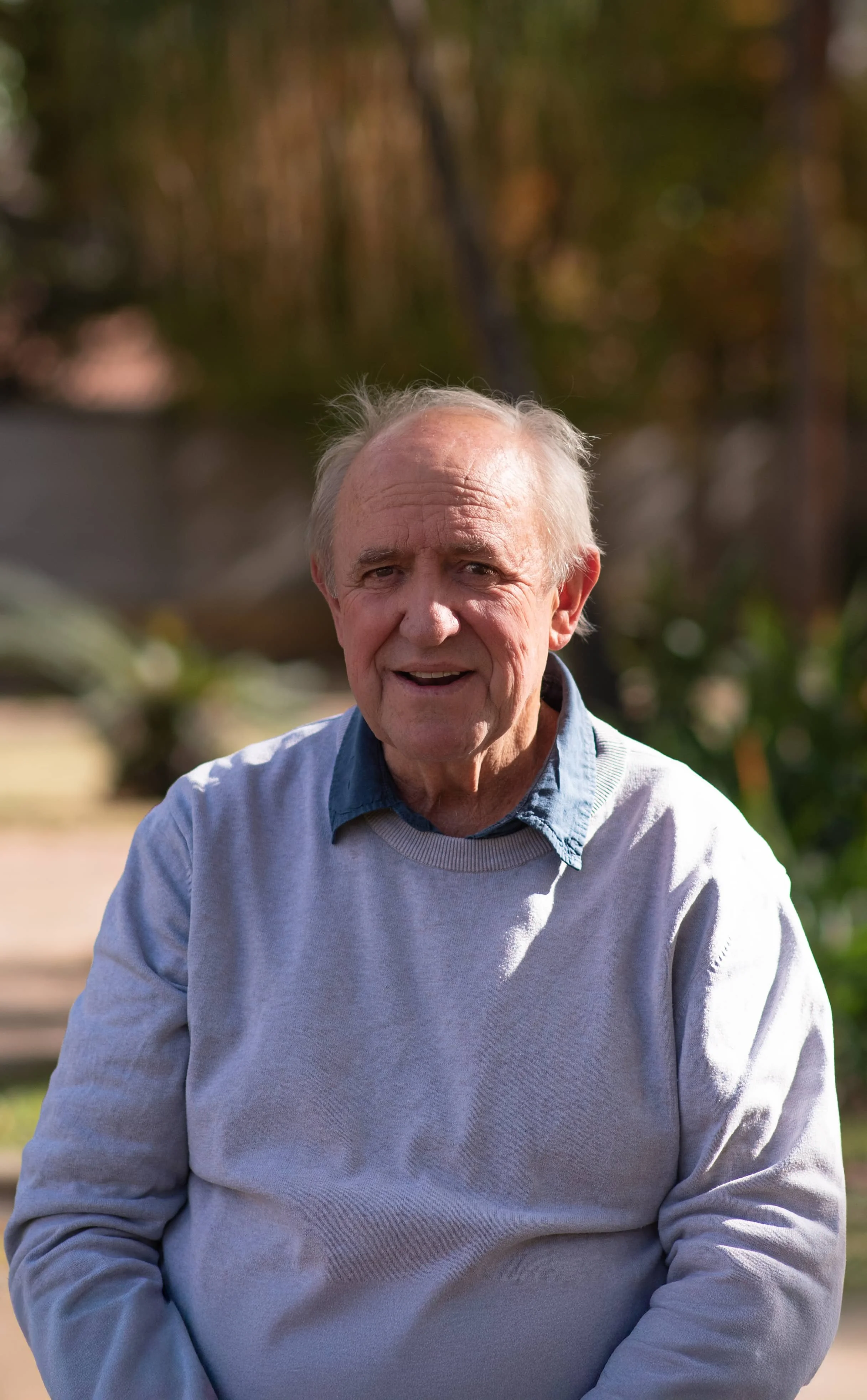 An elderly man with gray hair wearing a light gray sweater over a blue collared shirt, standing outdoors in a park with trees and blurred foliage in the background.