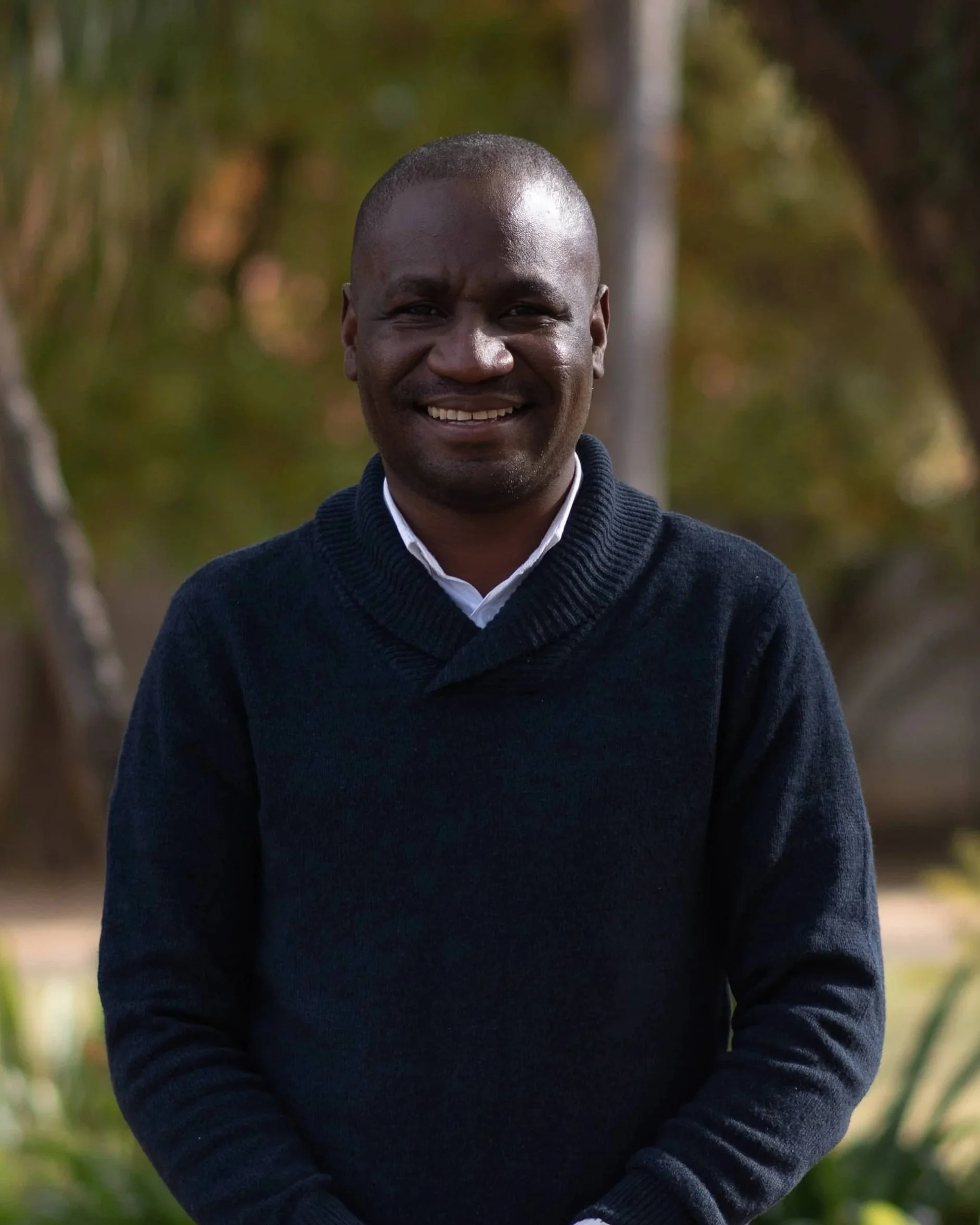 A smiling man with short hair, wearing a dark sweater over a collared shirt, standing outdoors with trees and greenery in the background.
