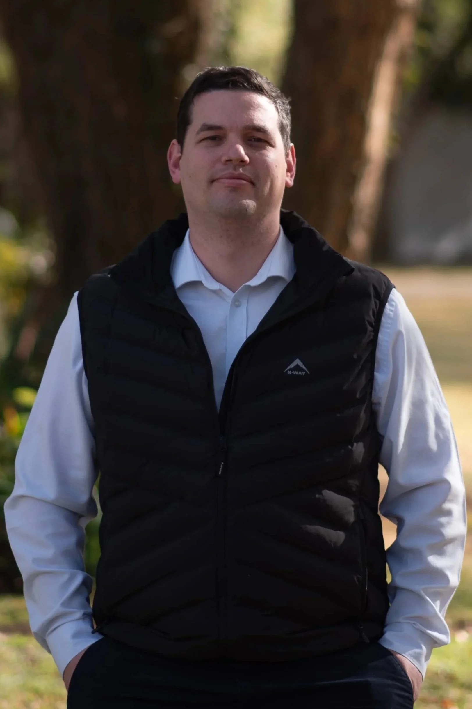 A man standing outdoors near a tree, wearing a white shirt and a black puffy vest, with hands in pockets.