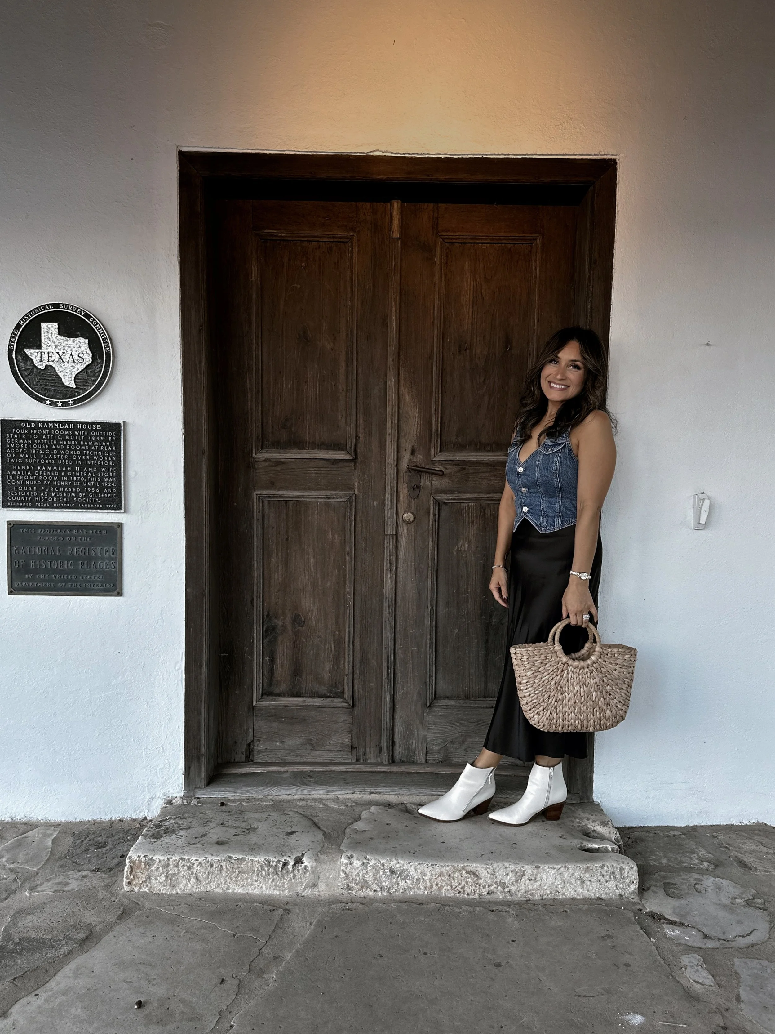 A woman standing in front of a wooden door holding a woven straw handbag, wearing a sleeveless denim vest, black skirt, white ankle boots, and smiling.