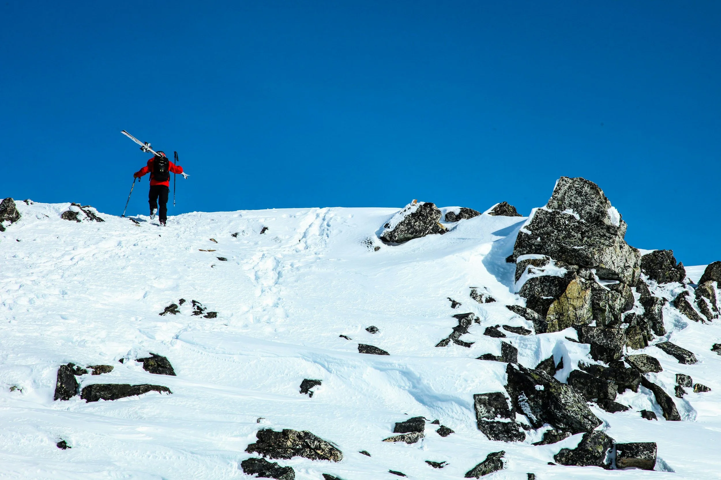 A person dressed in a red jacket carrying skis on their shoulder, walking up a snowy mountain slope with rocks, under a clear blue sky.