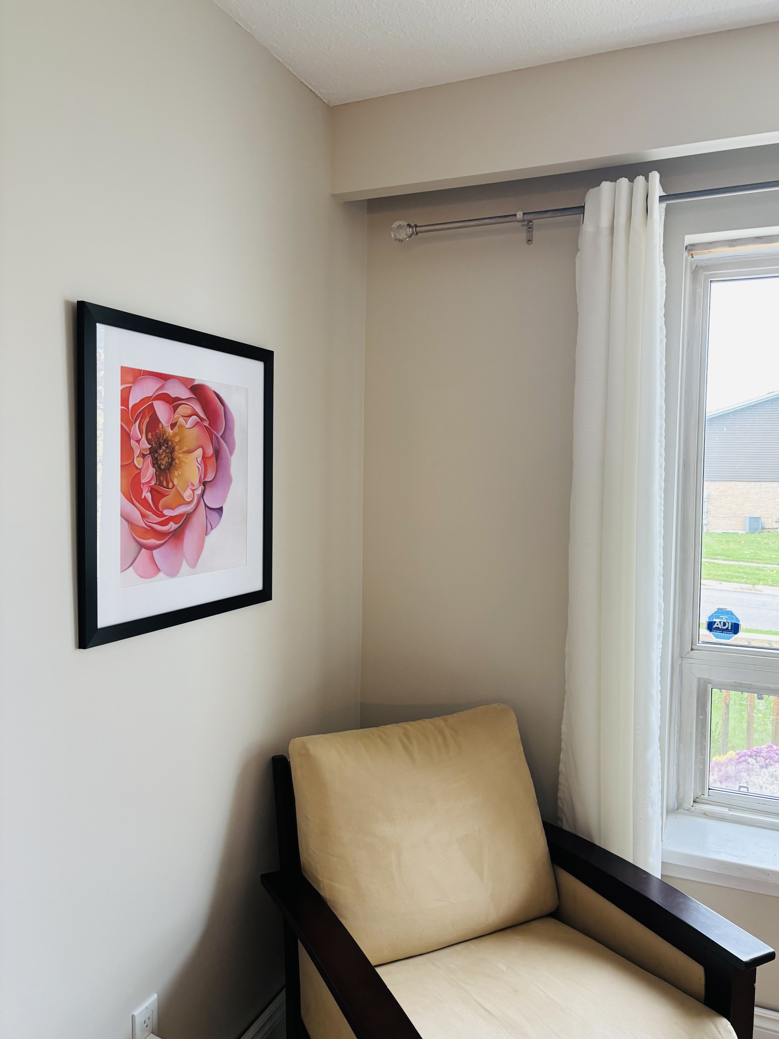 A beige armchair with dark wooden arms, a framed pink and orange flower artwork on the wall, a white curtain, and a window showing a grassy outdoor area.