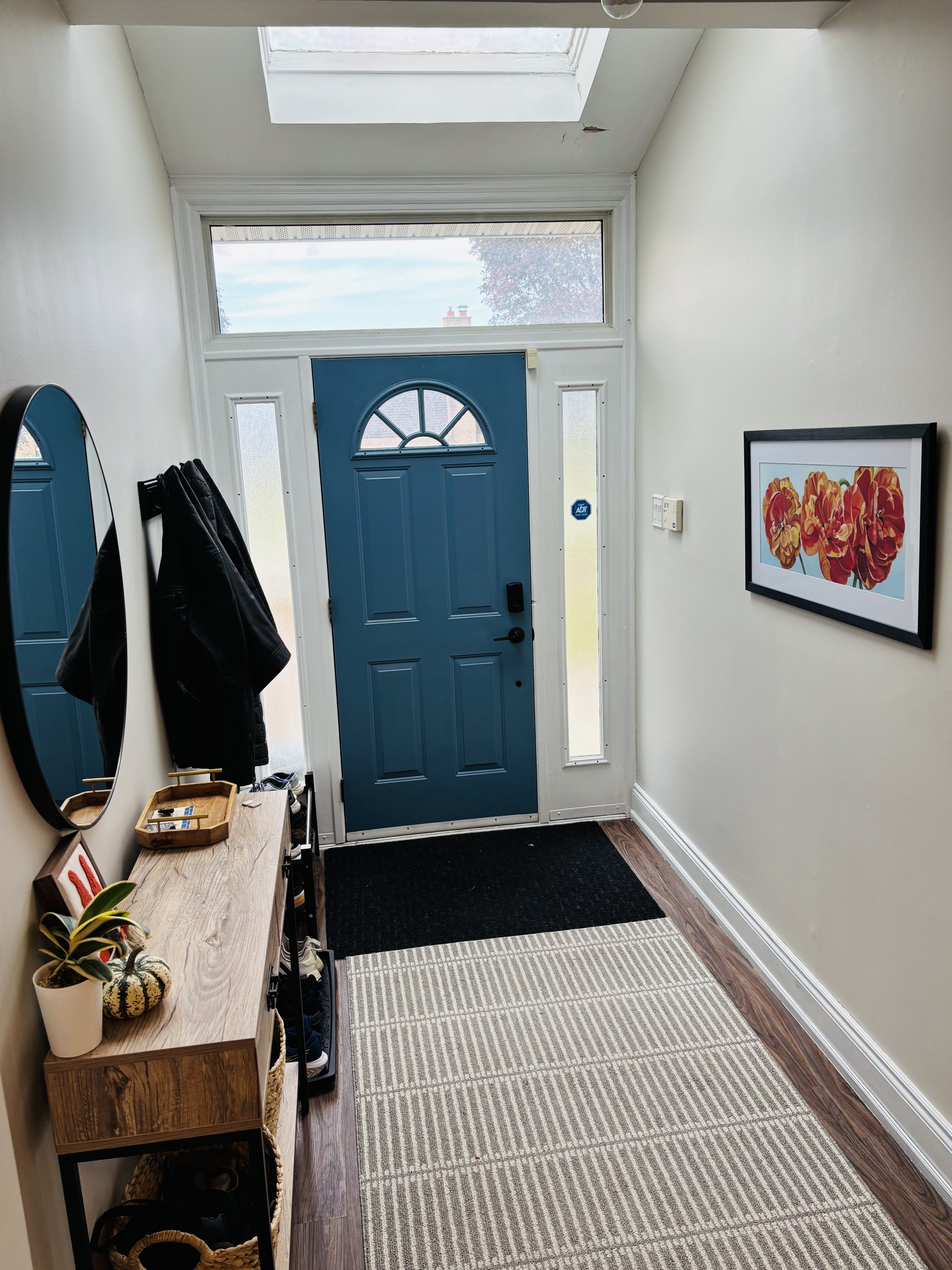 View of a home's front entryway with a blue door, side windows, a black door handle, a black doormat, a striped rug, a wooden console table with a potted plant, decorative items, and a framed floral artwork on the wall.