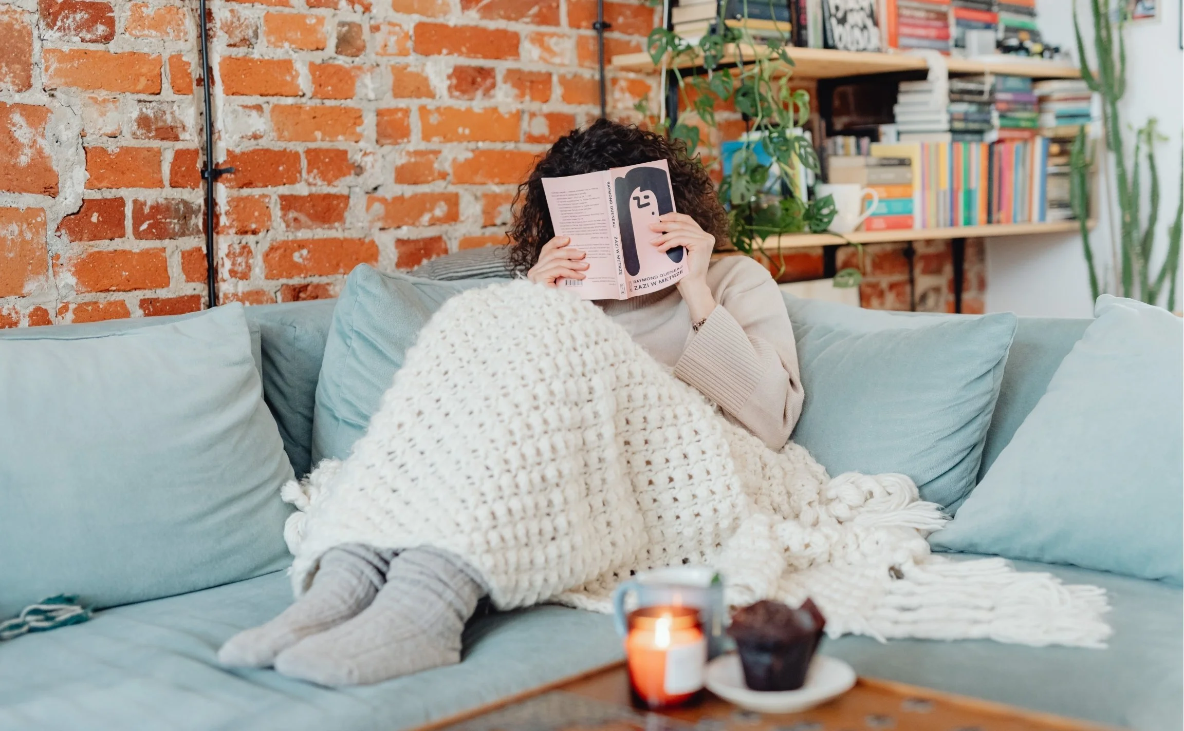 Person with curly hair reading a book while sitting on a blue sofa in a cozy room with brick walls, a wooden bookshelf filled with colorful books, and a large houseplant.