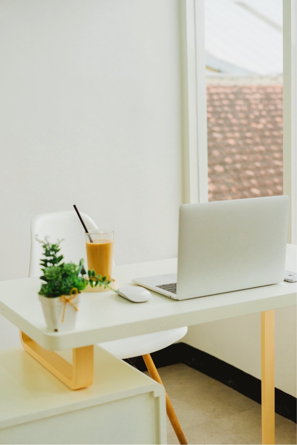 A minimalist workspace with a white desk and chair near a window. The desk has a laptop, a glass of iced tea with a straw, a white computer mouse, and a small potted plant. Outside the window, there is a view of a brick rooftop.