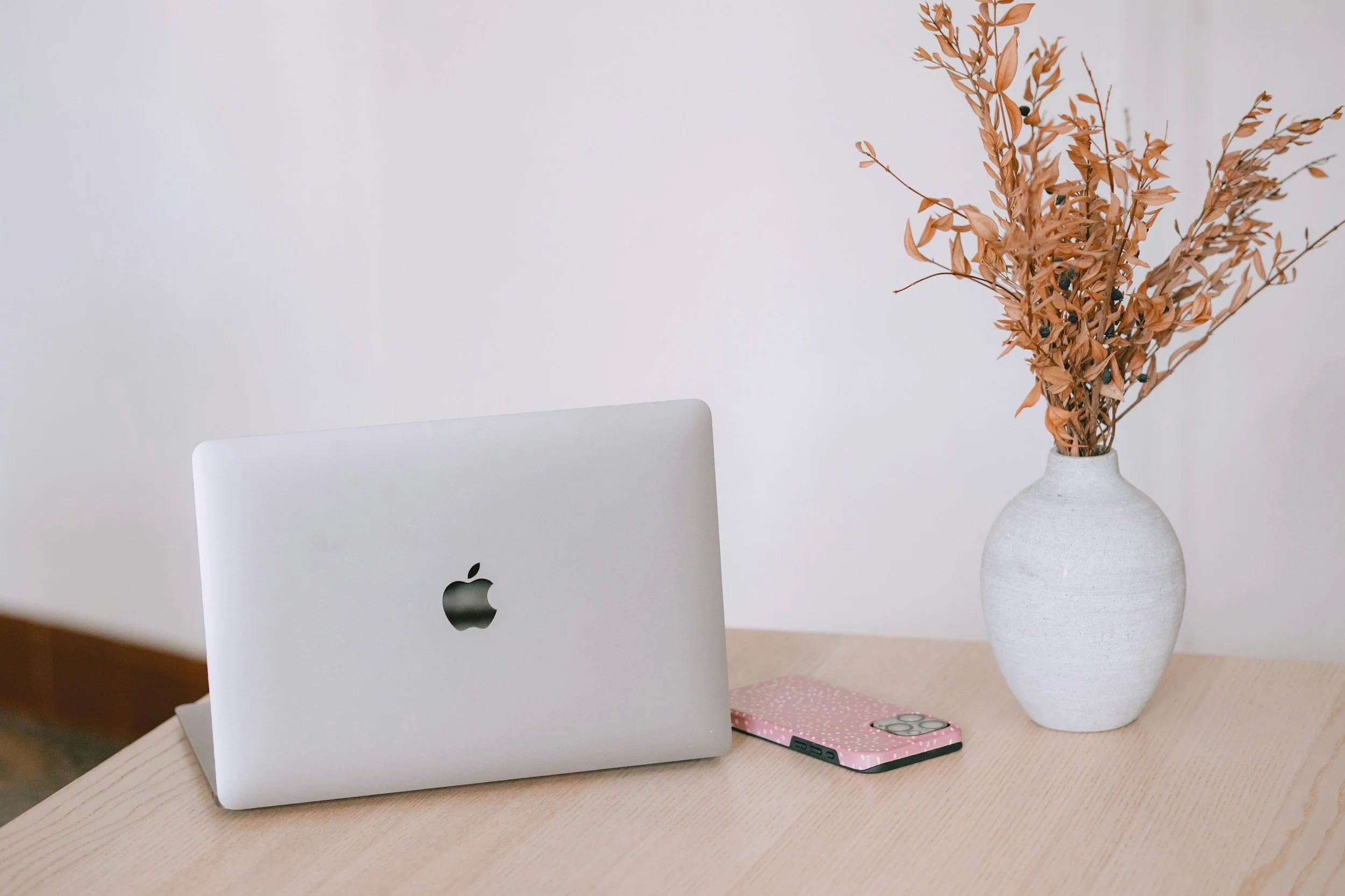 A desk with an Apple MacBook, a pink polka-dot phone case with an iPhone, a white ceramic vase with dried orange leaves, and a pink notebook on a light wood surface.