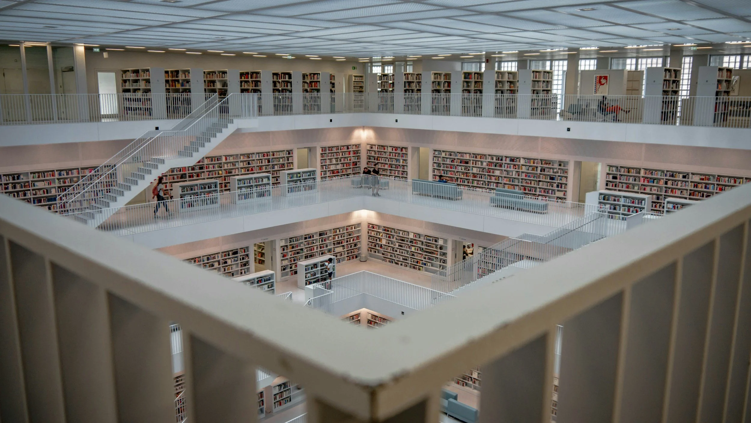 Multiple floors of a modern library with white railings, bookshelves filled with books, and a few people browsing.