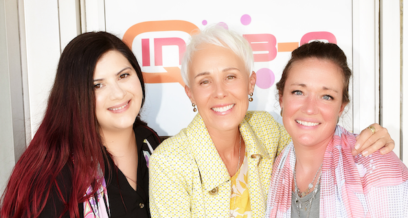 Three women smiling and posing together in front of a branded backdrop at an event.