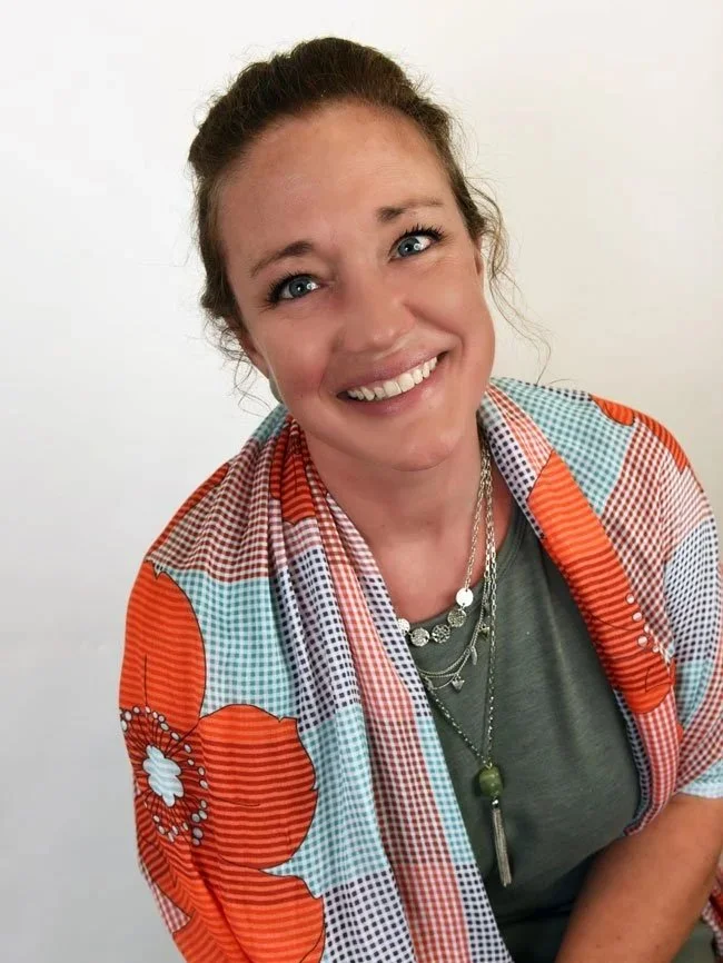 Smiling woman with blue eyes, brown hair in a bun, wearing layered necklaces and a colorful patterned kimono over a green shirt, standing against a white background.