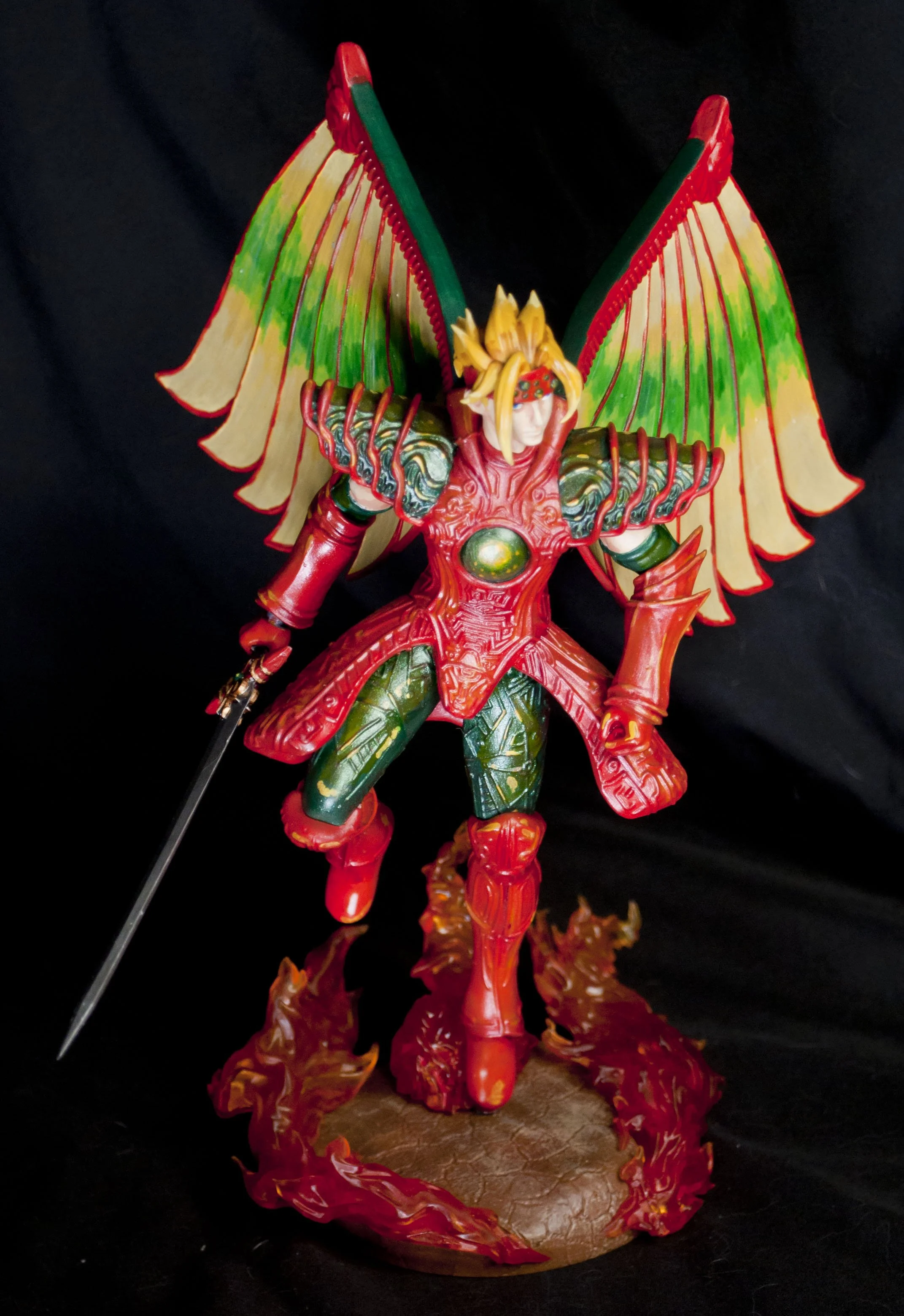 Colorful figurine of a fierce female warrior with large rainbow-colored wings, wearing red and green armor, holding a sword, with a fiery base underneath.