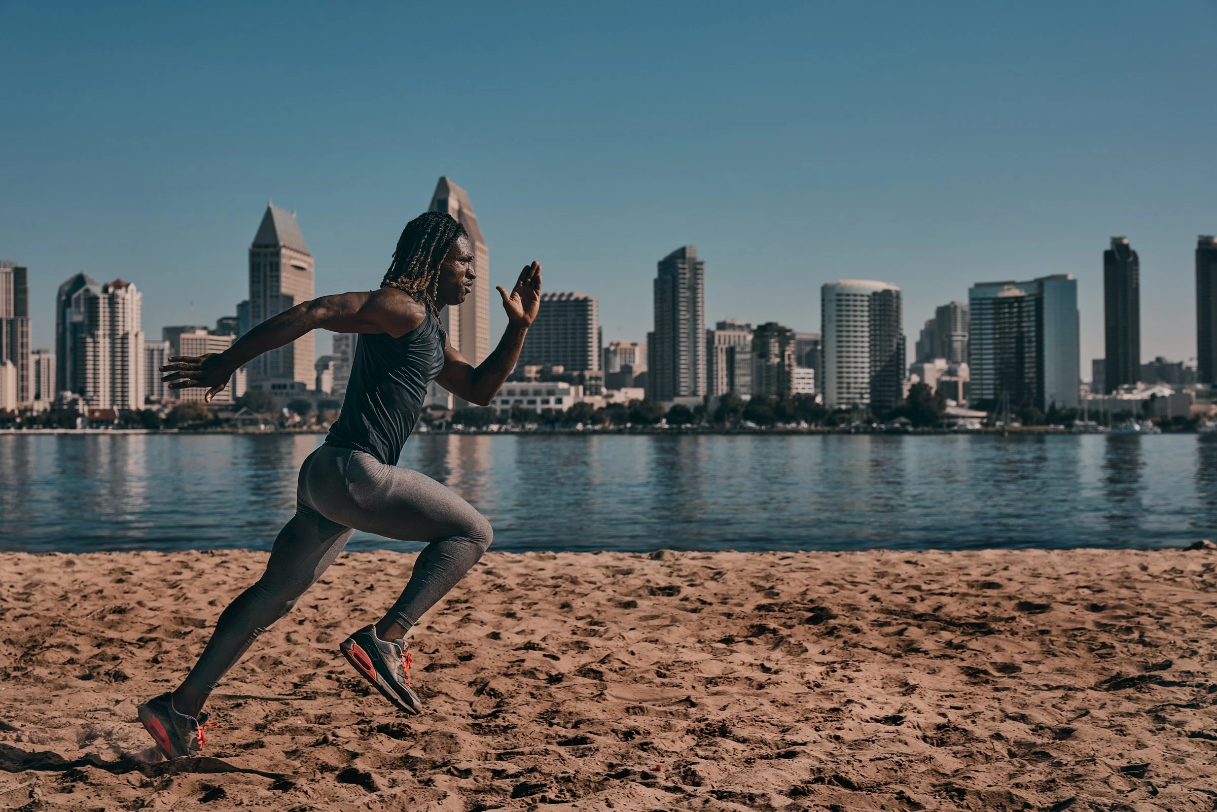 A man is running on the beach near water with a city skyline in the background.