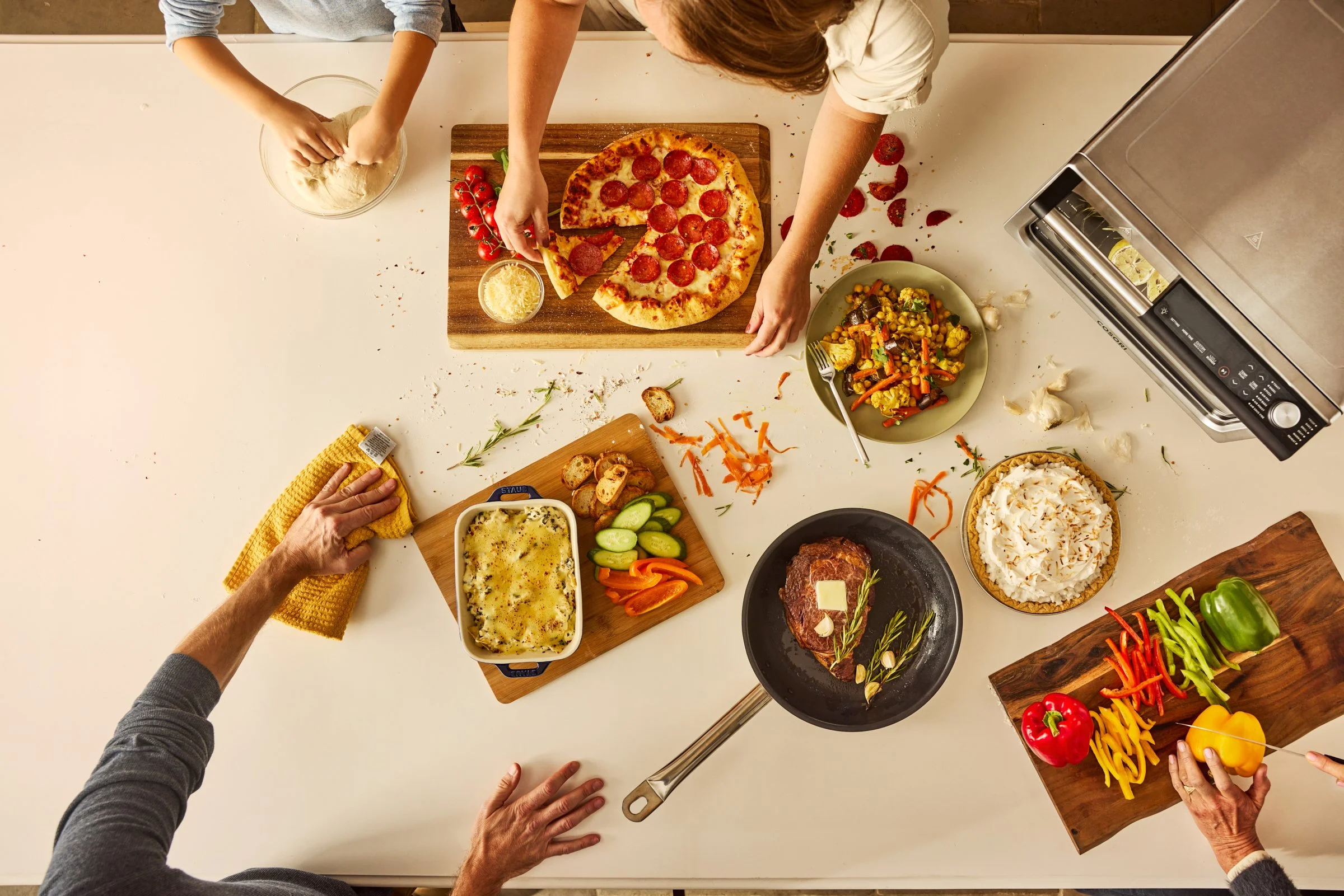 Top-down view of a family preparing a meal in the kitchen. There are various dishes on the table, including a pepperoni pizza on a wooden board, a bowl of salad, a casserole dish, a cast iron skillet with seasoned meat, a bowl in front with whipped c