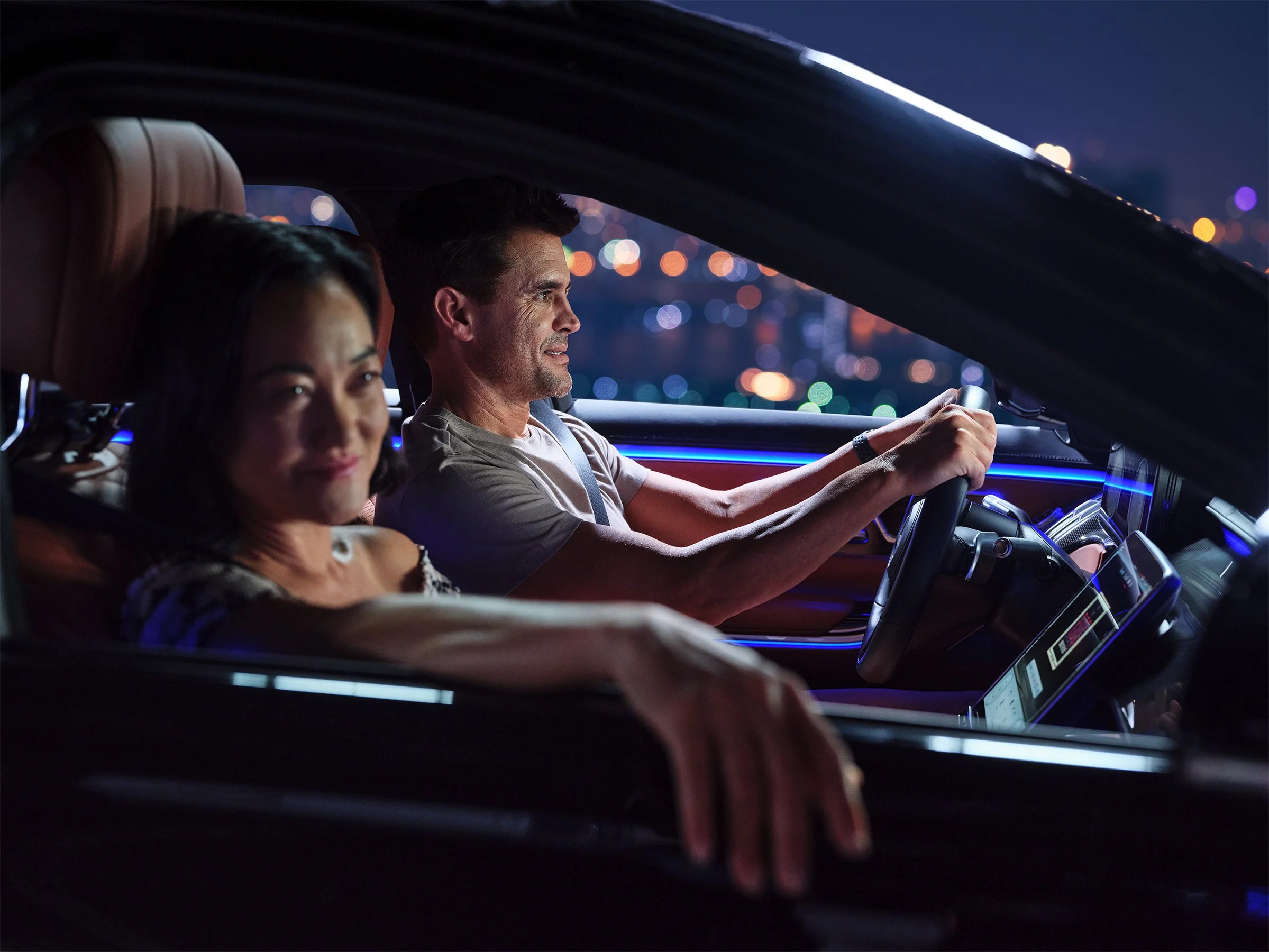 A man is driving a car at night with a woman sitting next to him. The city lights are visible in the background through the windows.