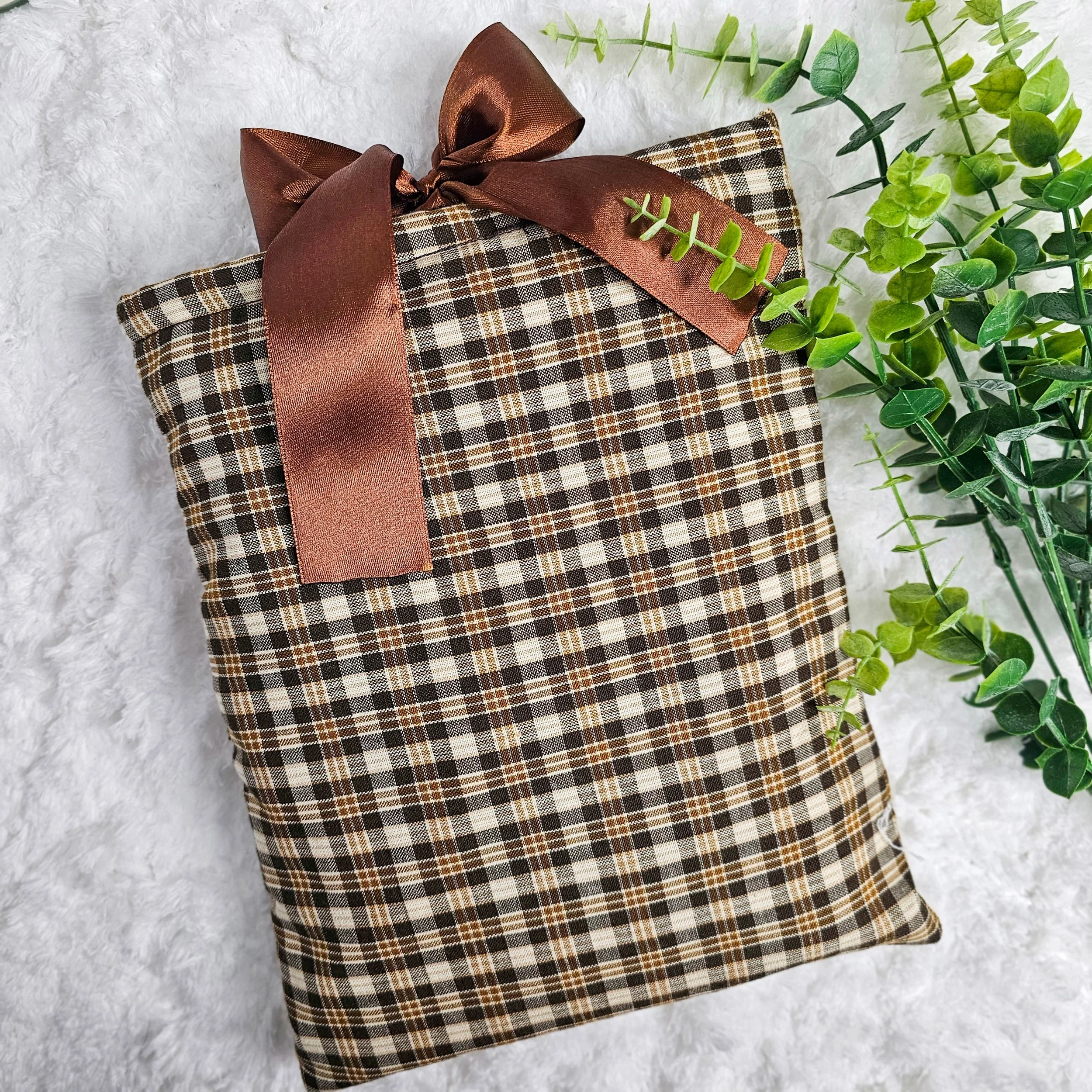 A wrapped gift with brown, beige, and white checkered patterned paper, adorned with a brown satin ribbon tied into a bow, placed on a white fluffy surface next to green leafy plants.