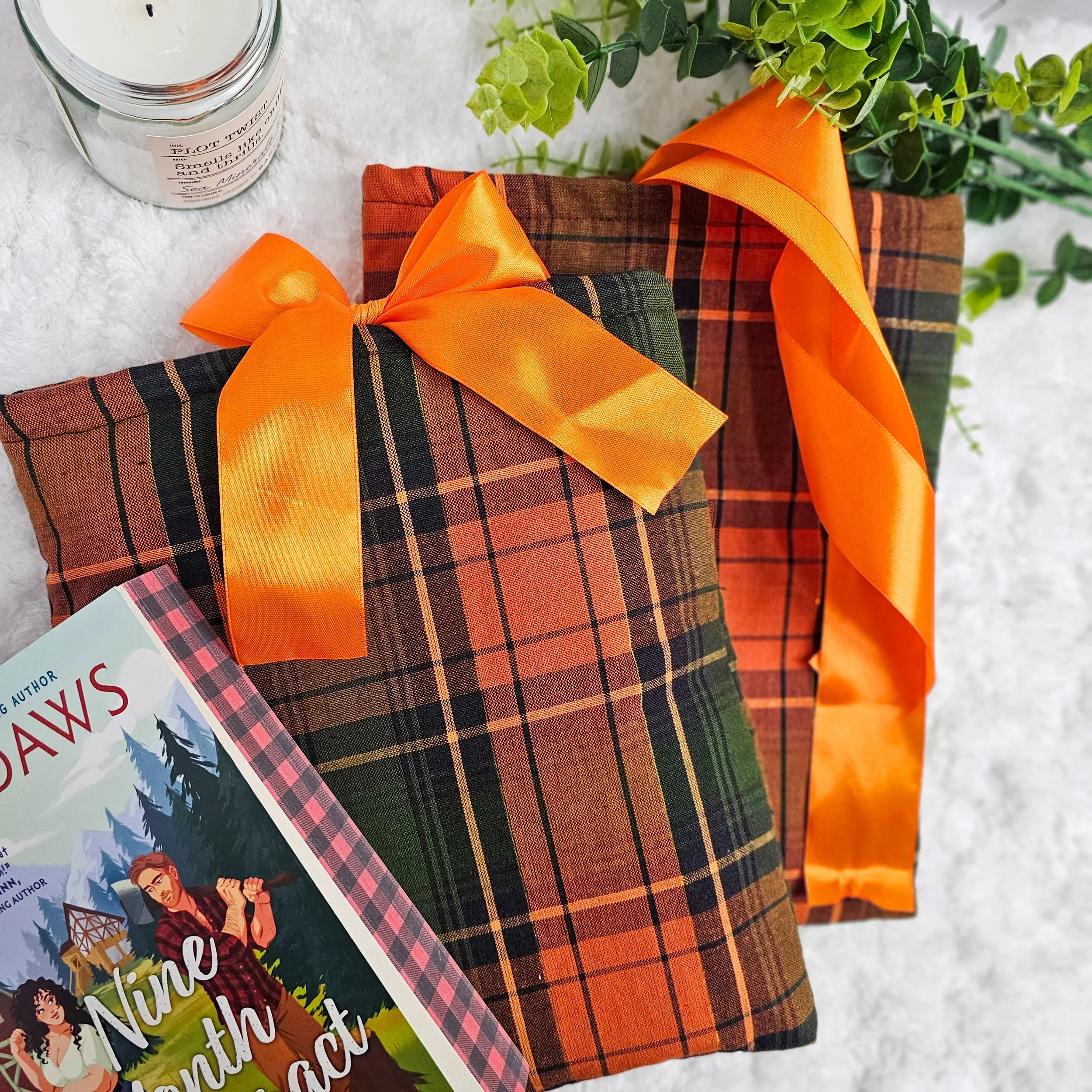 Two wrapped gifts with orange satin ribbons, a book titled 'Nine Anti Hack' partially visible, a scented candle, and green leafy plants on a white textured surface.