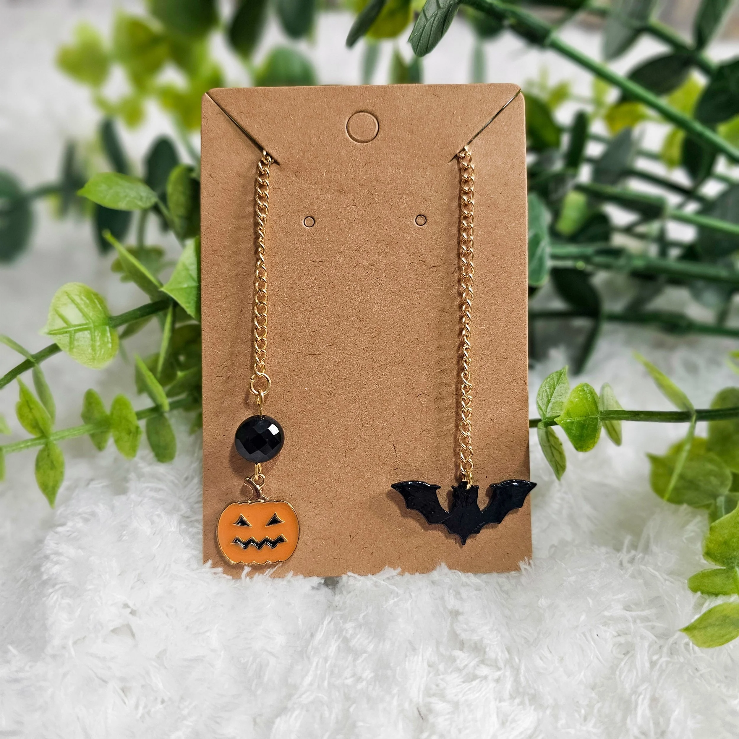 Two Halloween-themed earrings on a brown display card with green foliage and white fluffy background. The left earring has a orange pumpkin charm with a black face and a black bead above it, the right earring has a black bat-shaped charm.