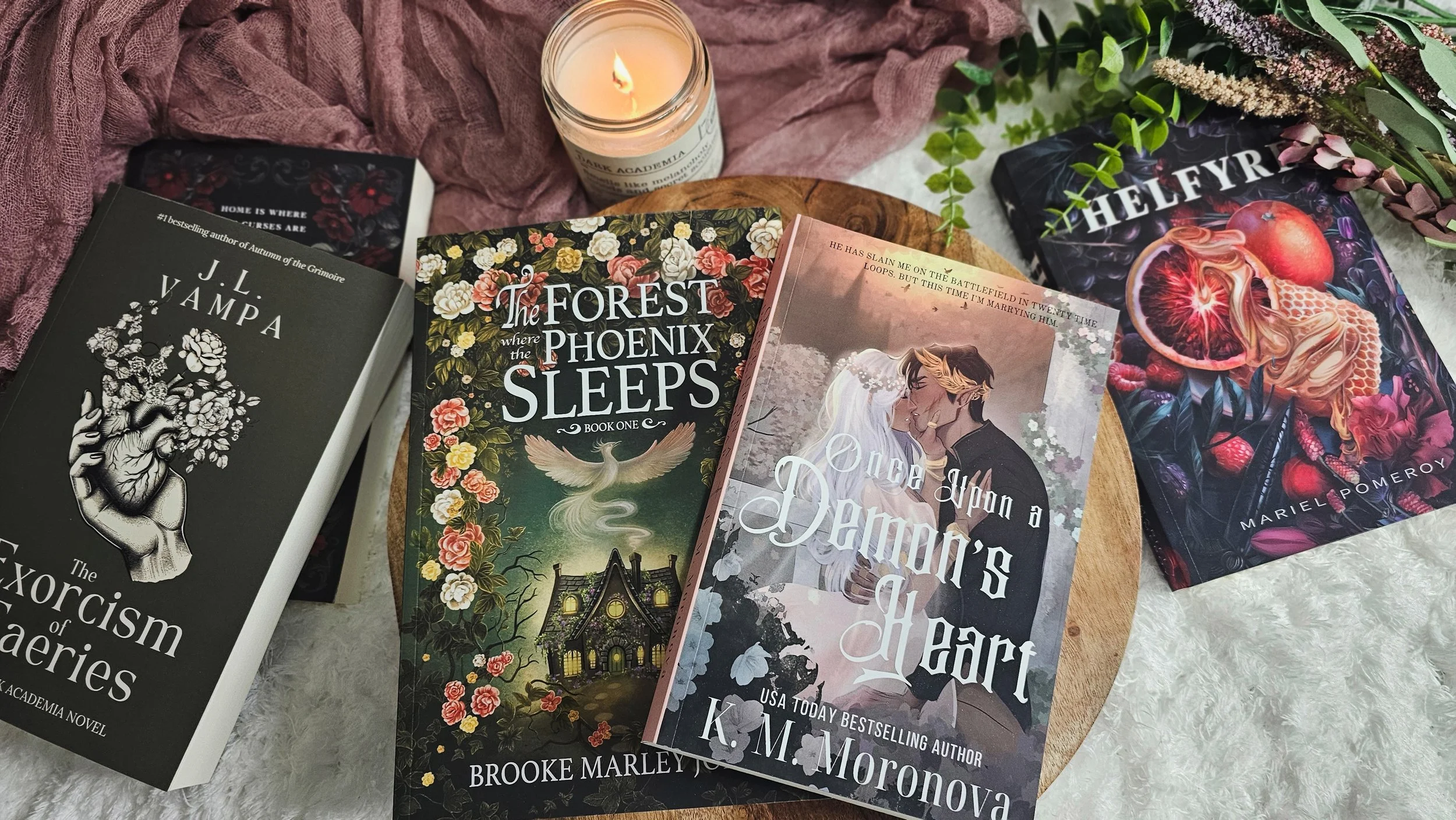 A collection of five books with fantasy and romance themes, placed on a wooden table with a lit candle and greenery in the background.