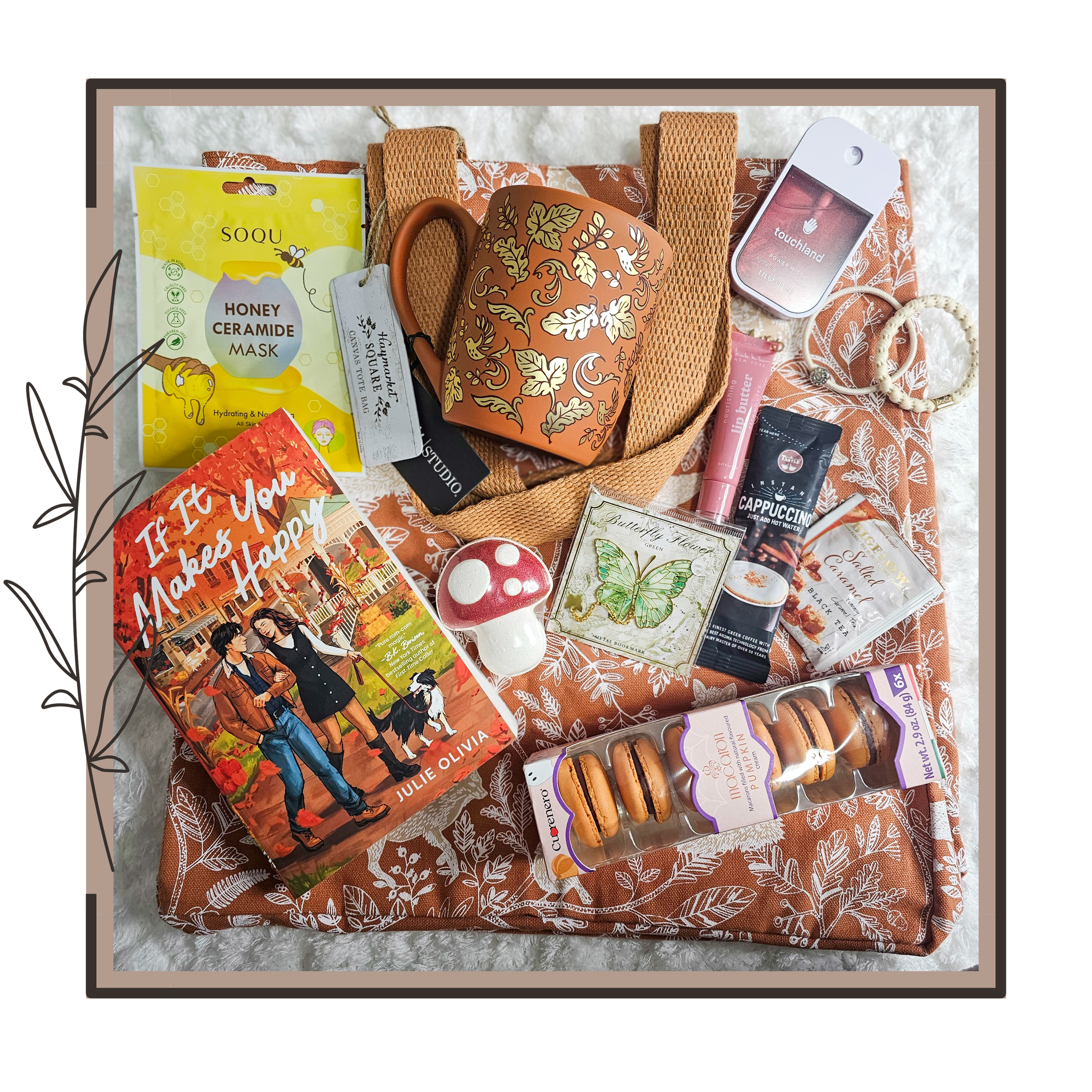 A collection of personal items and gifts arranged on a brown floral-patterned tote bag. Includes a paperback book titled "If It Makes You Happy," a potted cactus-shaped decor, a mushroom-shaped ceramic item, a pack of macarons, a honey ceramide face mask, a mug with an ornate floral design, a pack of green tea, a tube of lip balm, a bag of cappuccino, a Bali-inspired beaded bracelet, a white beaded hair tie, a pack of skin scrub, and a small electronic device, all laid on a white fabric background.