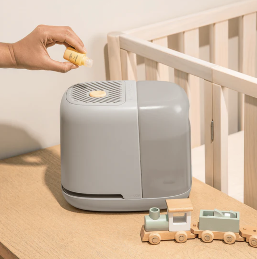 A hand is pouring a capsule into a gray portable humidifier or diffuser on a wooden table, with a wooden toy train and a crib in the background.