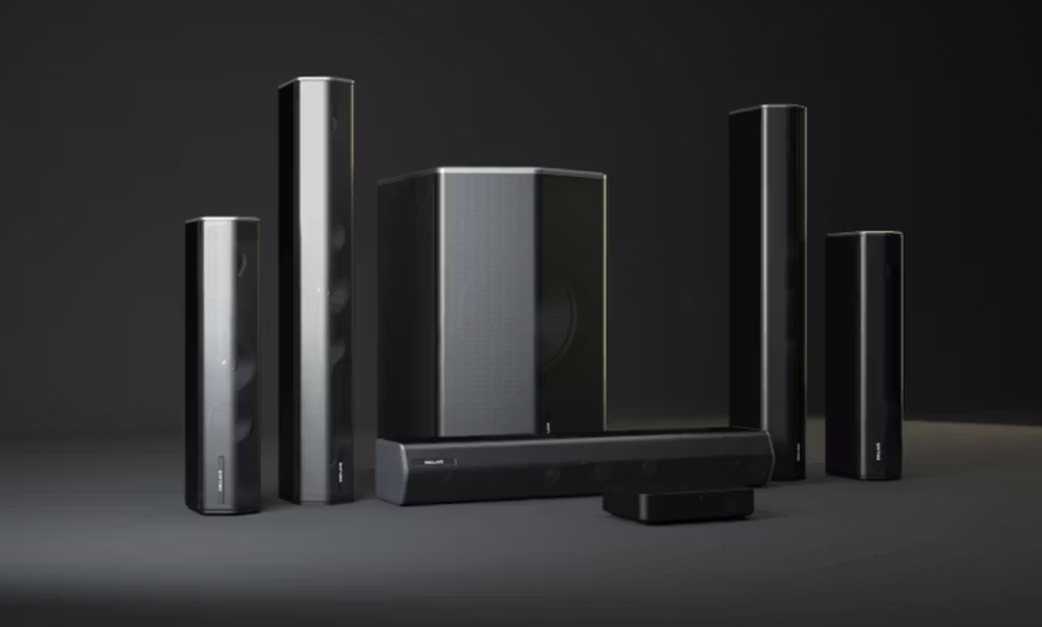 A home theater system with multiple black and silver speakers arranged in a row, including a central subwoofer and soundbar, on a dark surface against a dark background.