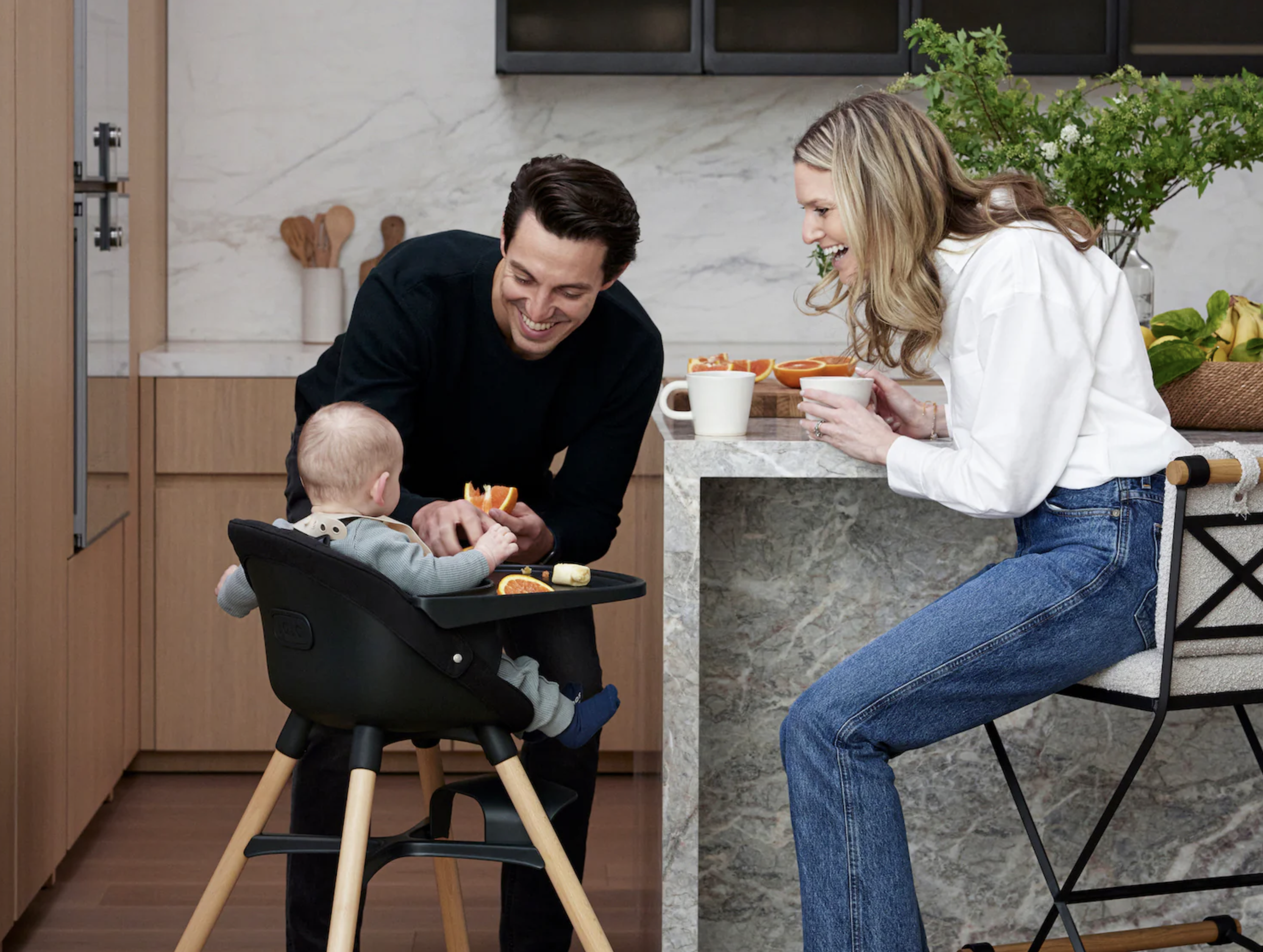 A family in a kitchen with a man, woman, and a baby. The man is smiling and leaning over a baby in a high chair, holding an orange slice. The woman is sitting on a chair, leaning on a marble counter, smiling, and holding a white mug. There are oranges on the counter and in mugs, with a vase of flowers in the background.