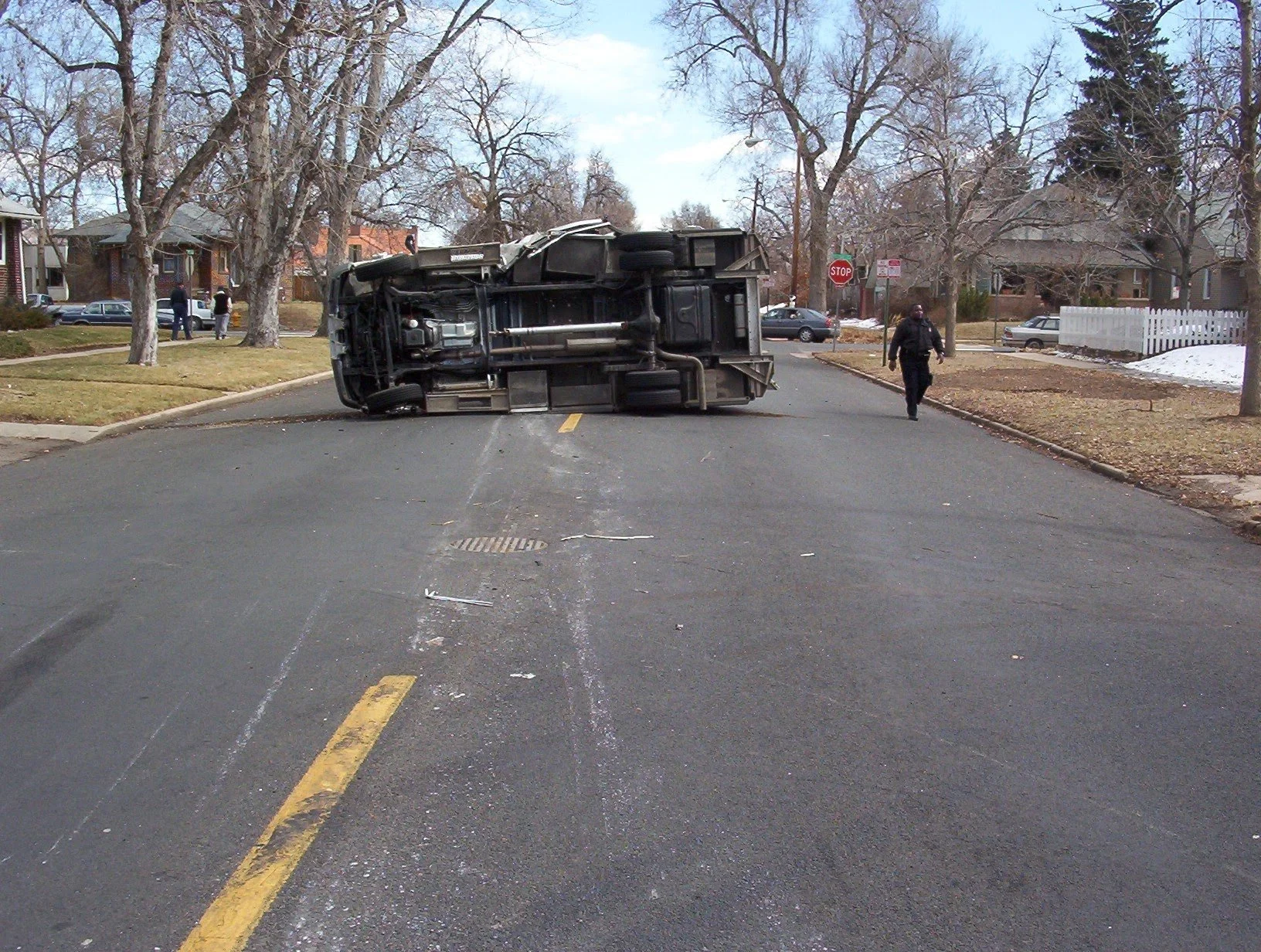 A flipped-over vehicle blocking a residential street with people and police officers nearby.
