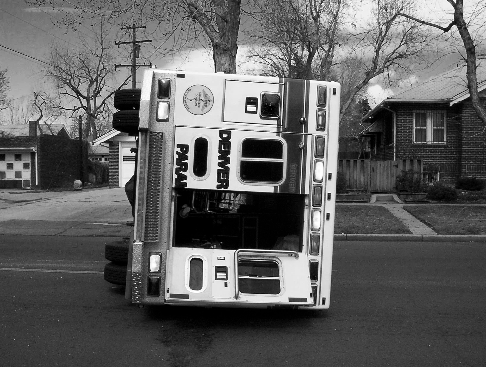 An ambulance truck flipped onto its side on a residential street in black and white.