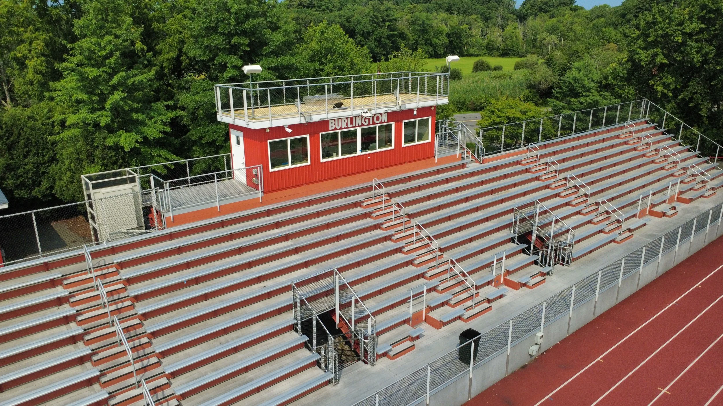 Aerial view of a red and white metal bleacher seating area at a sports stadium with a small press box labeled 'BURLINGTON' at the top, surrounded by green trees.