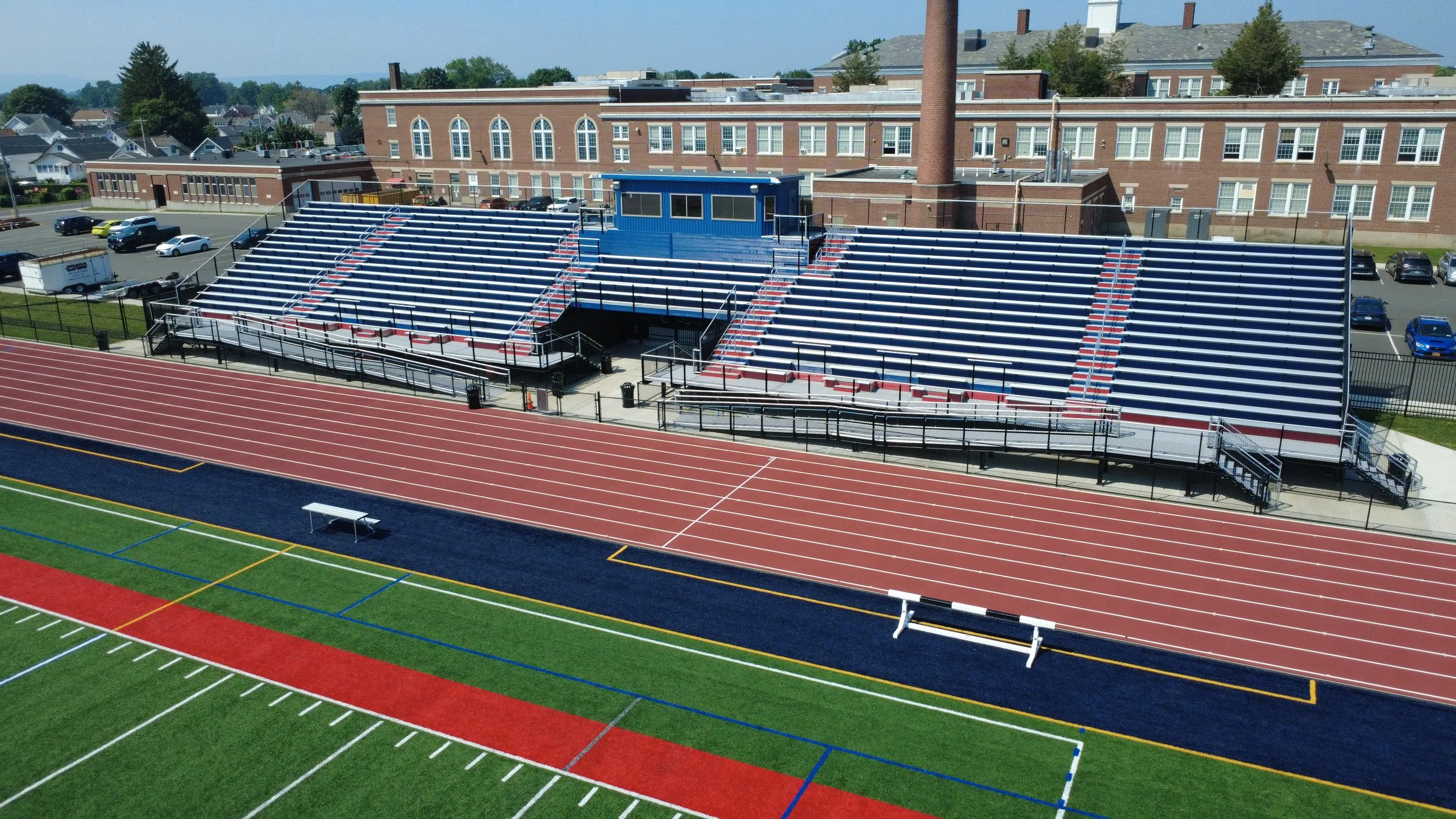 An outdoor sports stadium with bleachers and a running track, adjacent to a school building with brick walls and multiple windows. The bleachers are empty, and parking lots with cars are visible behind the bleachers.