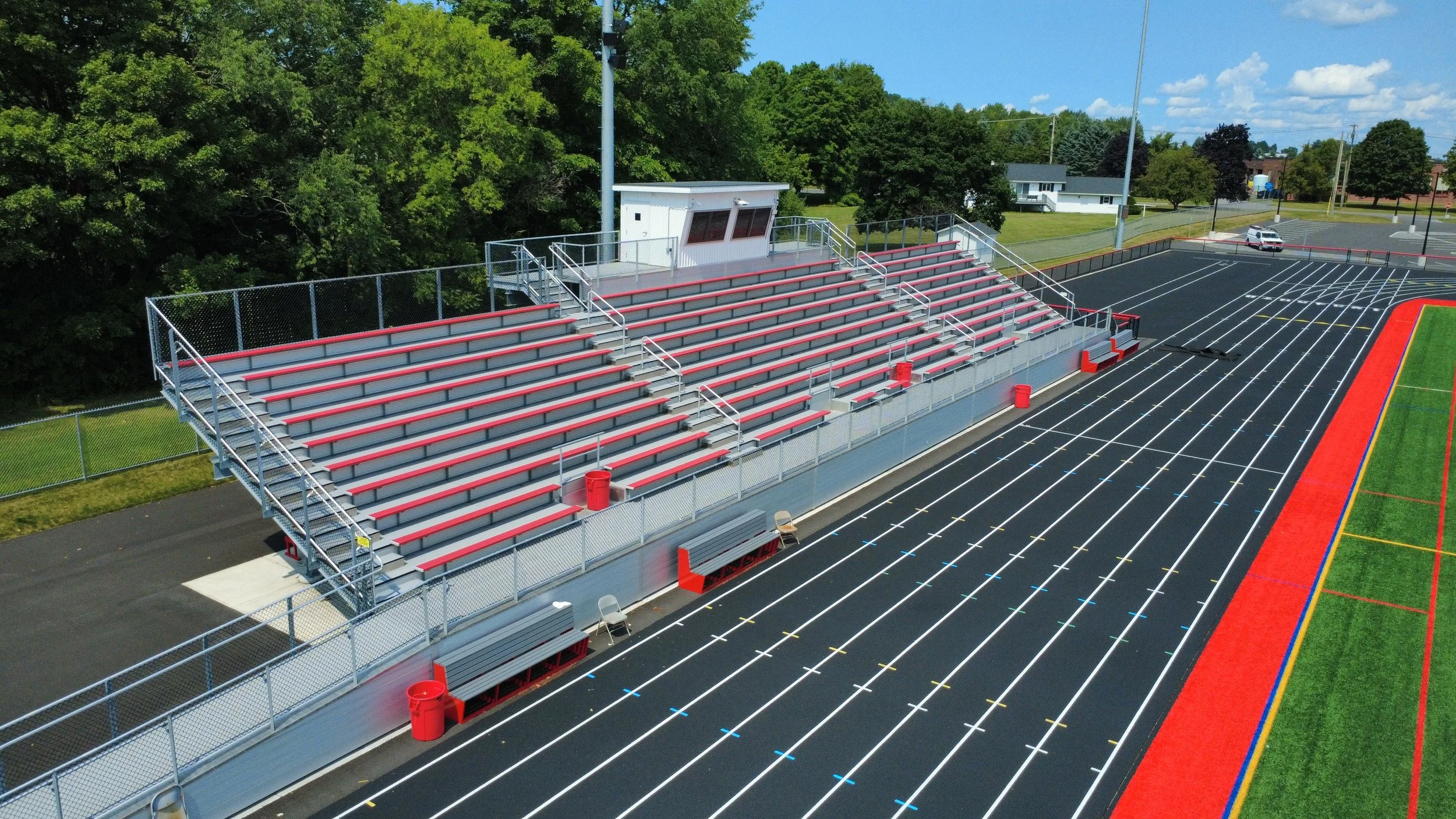 Empty bleachers and a track at a sports stadium next to trees and houses, under a partly cloudy sky.