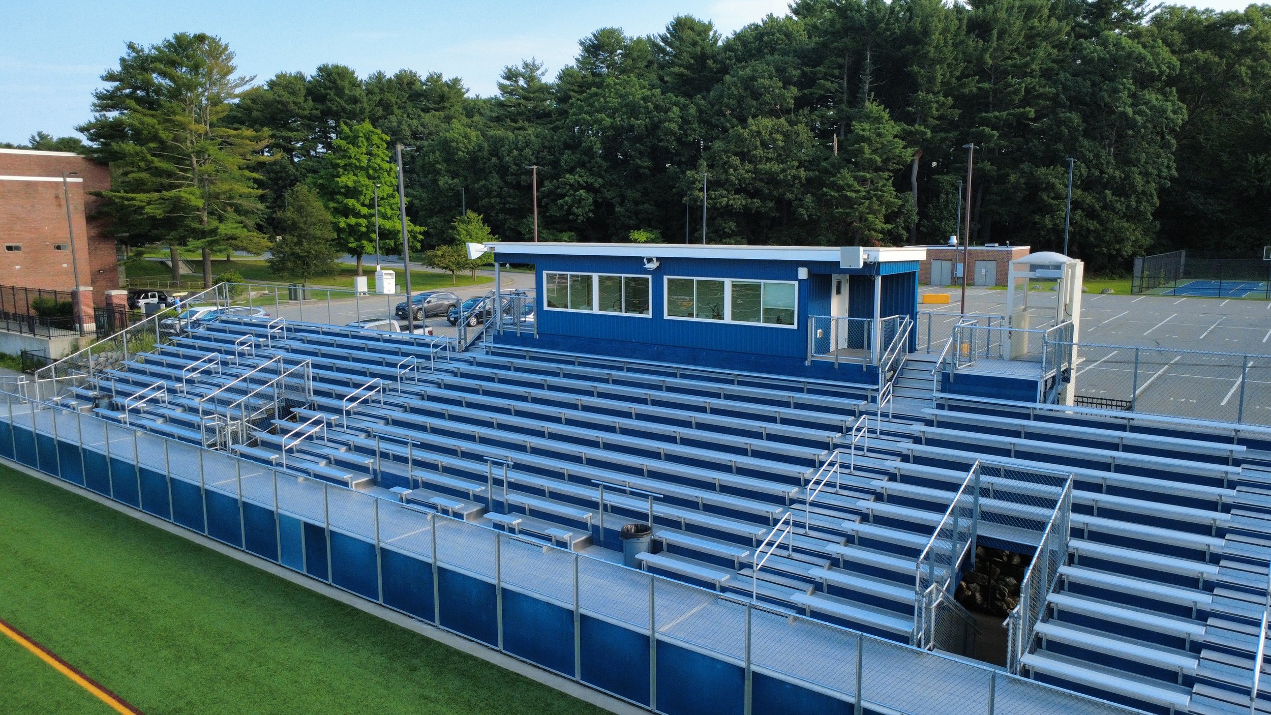 Empty blue bleachers at a sports field with a small blue building, parking lot and trees in the background.
