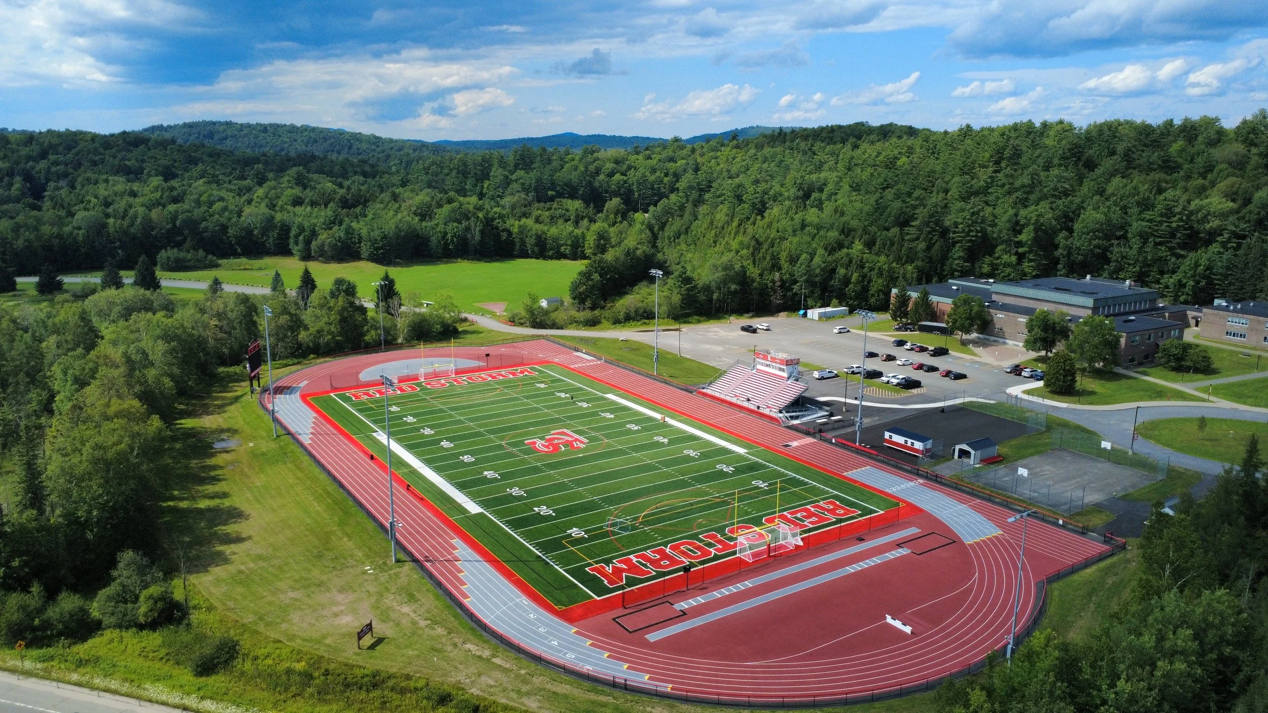 An aerial view of a football field and track with the words 'PEEL COUNTY' and 'WELDOR' painted on the field, surrounded by a parking lot, fields, and a wooded area.